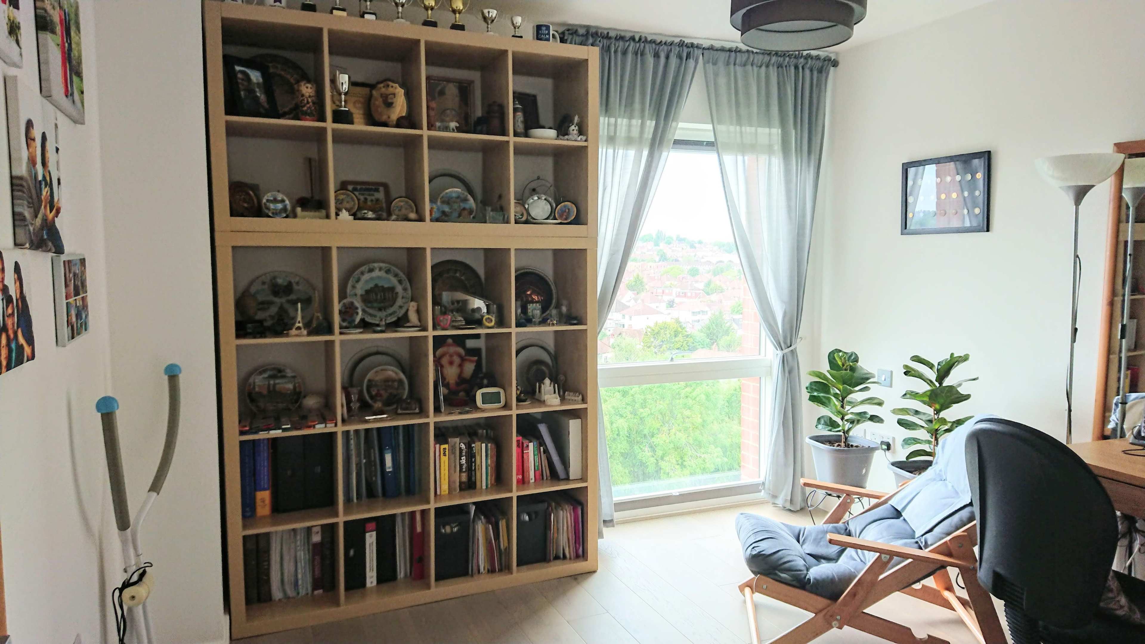 A room featuring a bookshelf filled with trophies and decorative plates, a chair, and a large window with a view of greenery outside.