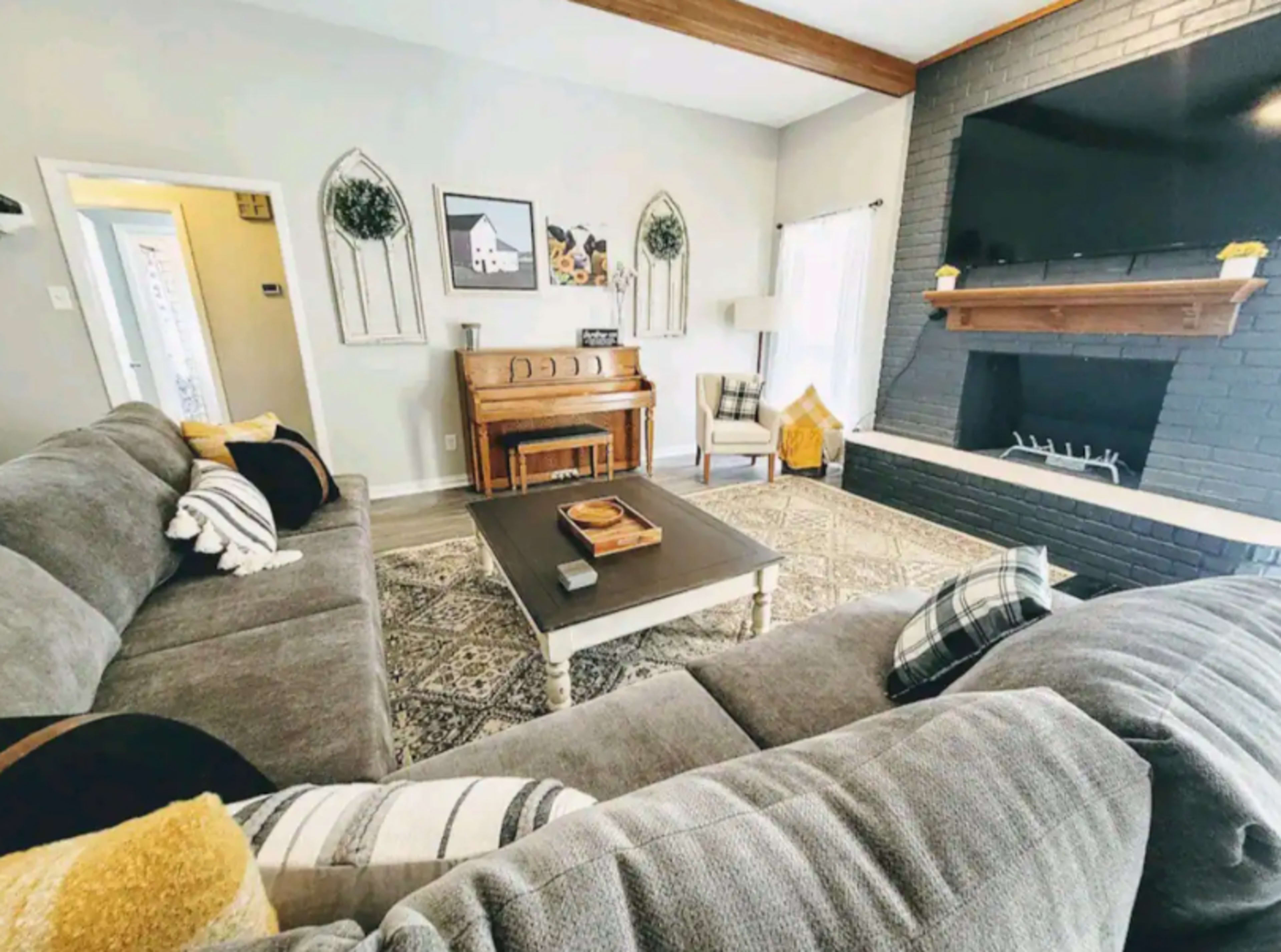 The living room features a gray sectional sofa, a wooden piano, and a black fireplace with a large TV mounted above it.