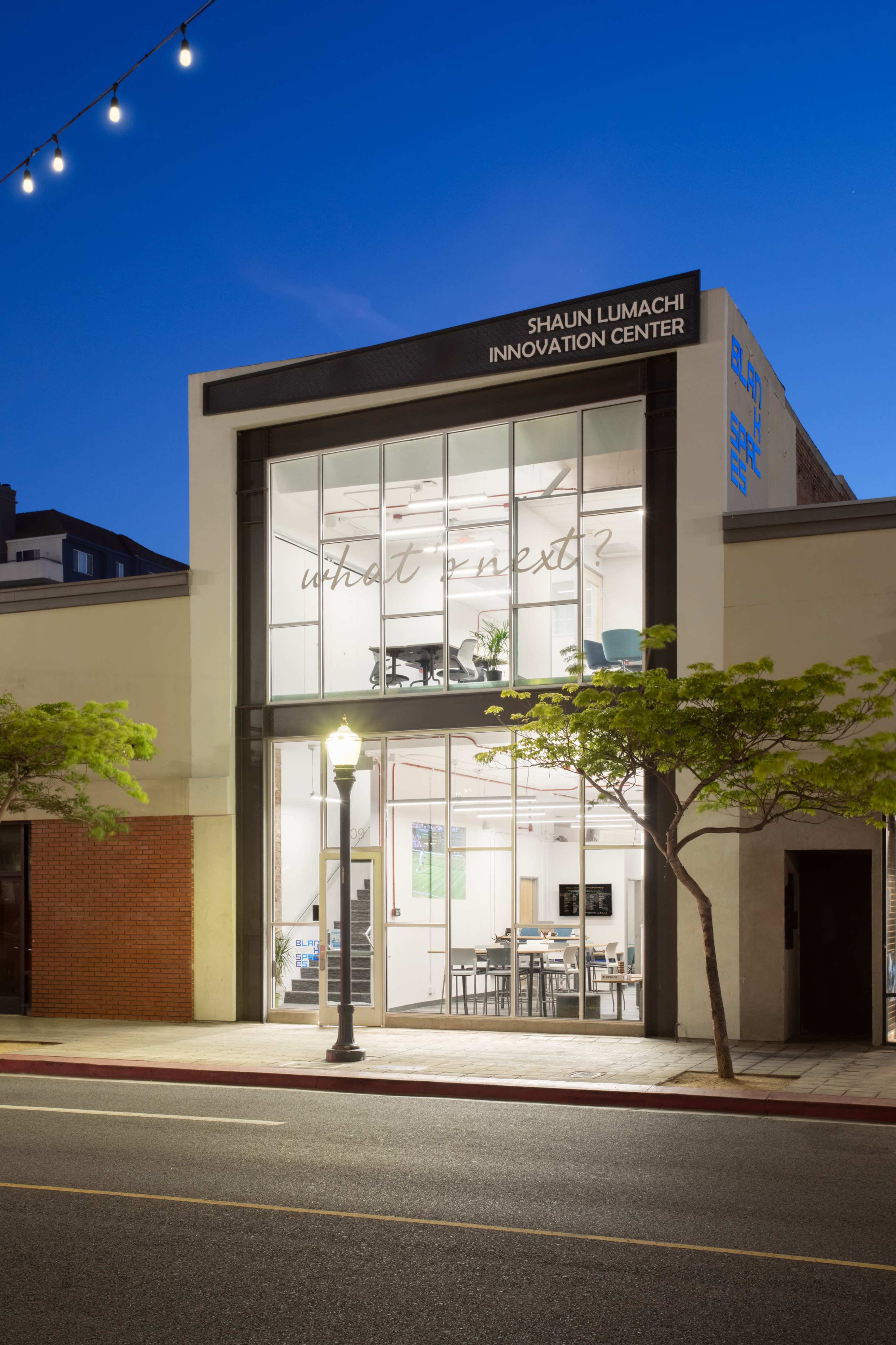 The exterior of the Shaun Lumachi Innovation Center, featuring large glass windows illuminated at dusk and a street lamp in front.