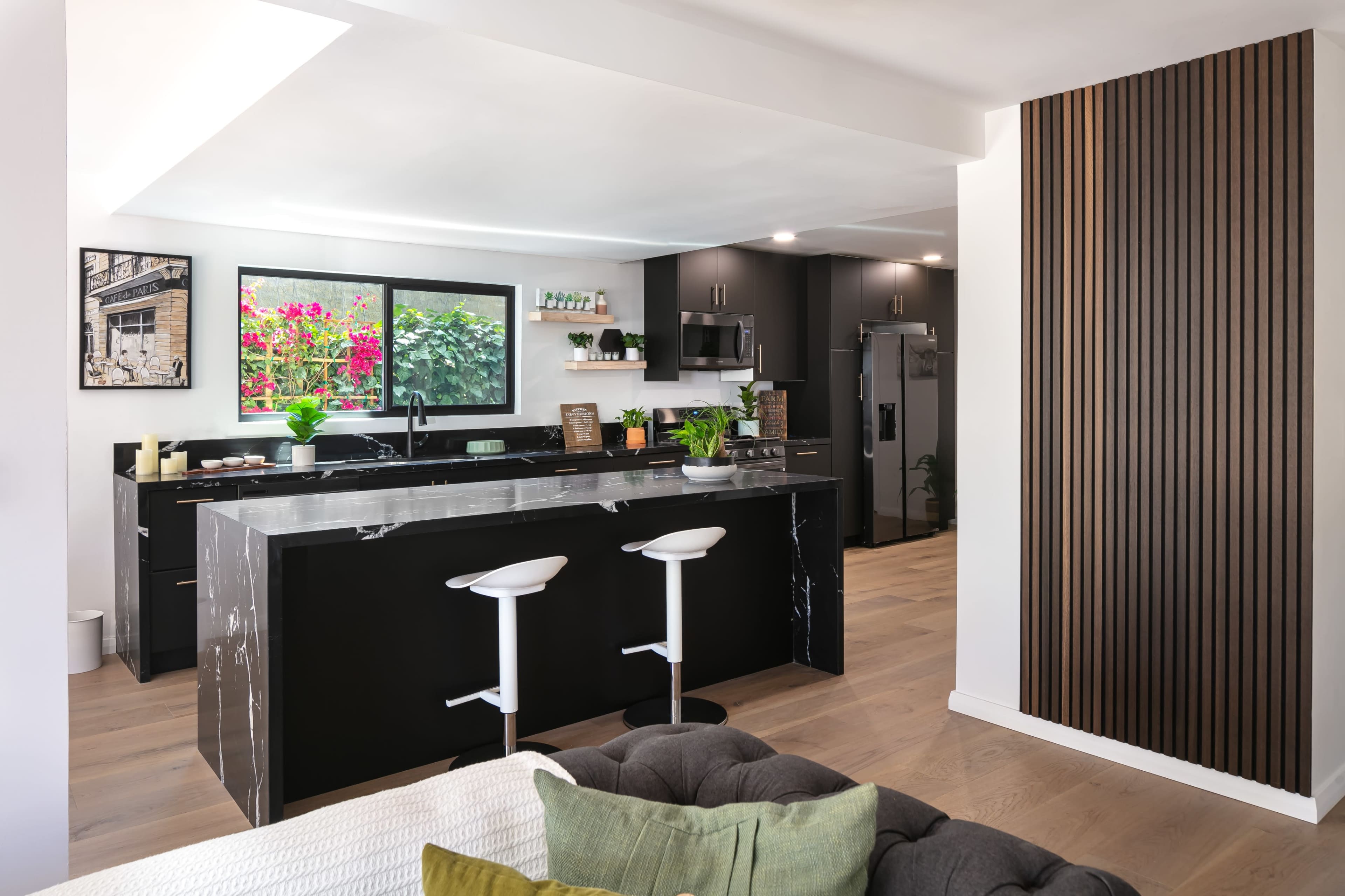 The image shows a modern kitchen featuring dark cabinetry, a marble countertop, and two white bar stools, with plants and flowers visible outside a large window.