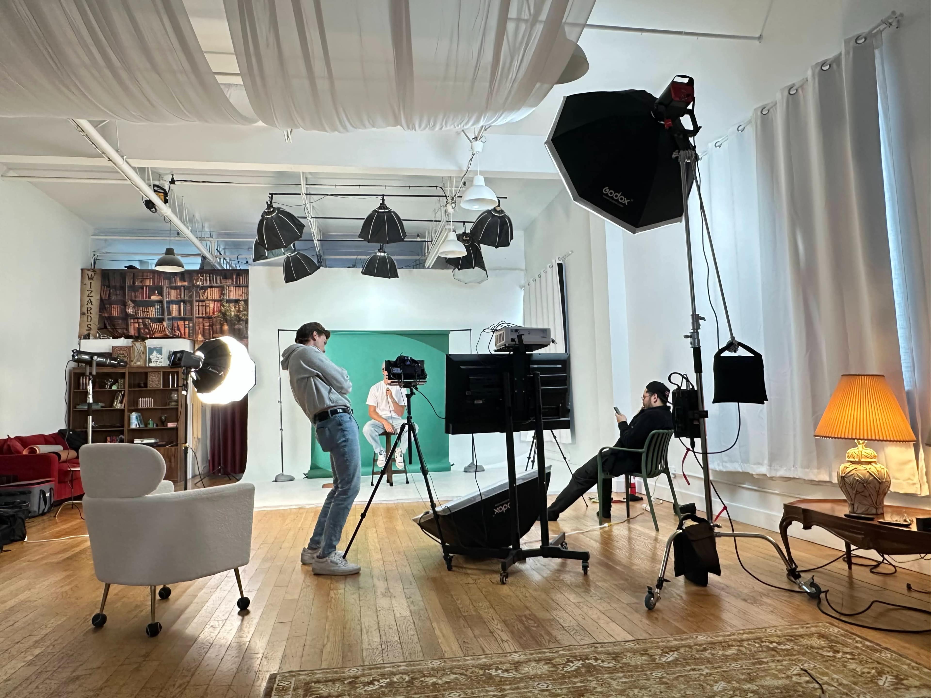 A film crew sets up in a studio featuring a green screen, lighting equipment, and various props, as two people prepare for a shoot.
