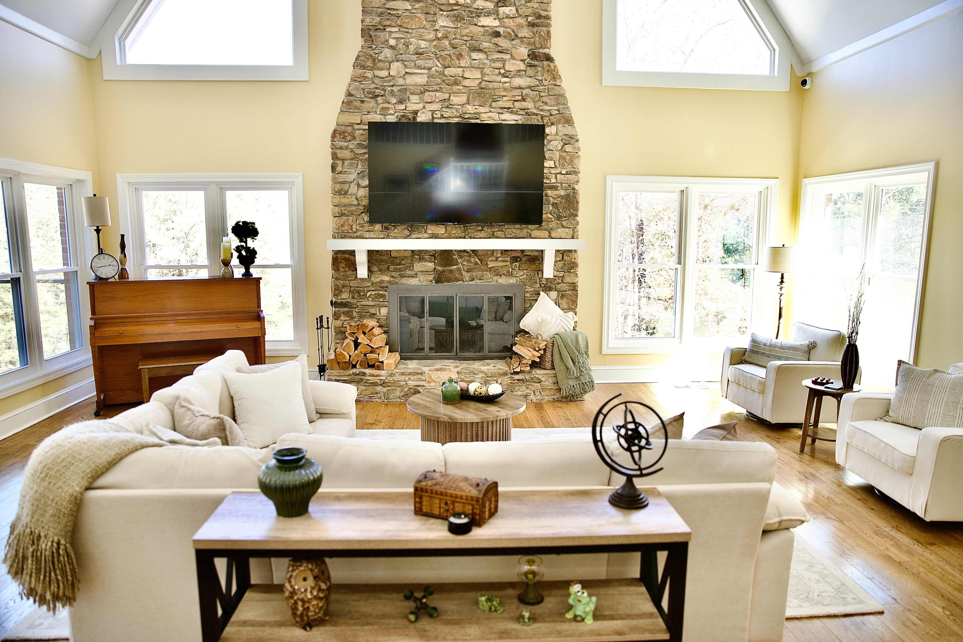 The image features a spacious living room with a stone fireplace, a large television mounted above it, and comfortable seating arrangements surrounding a wooden coffee table.