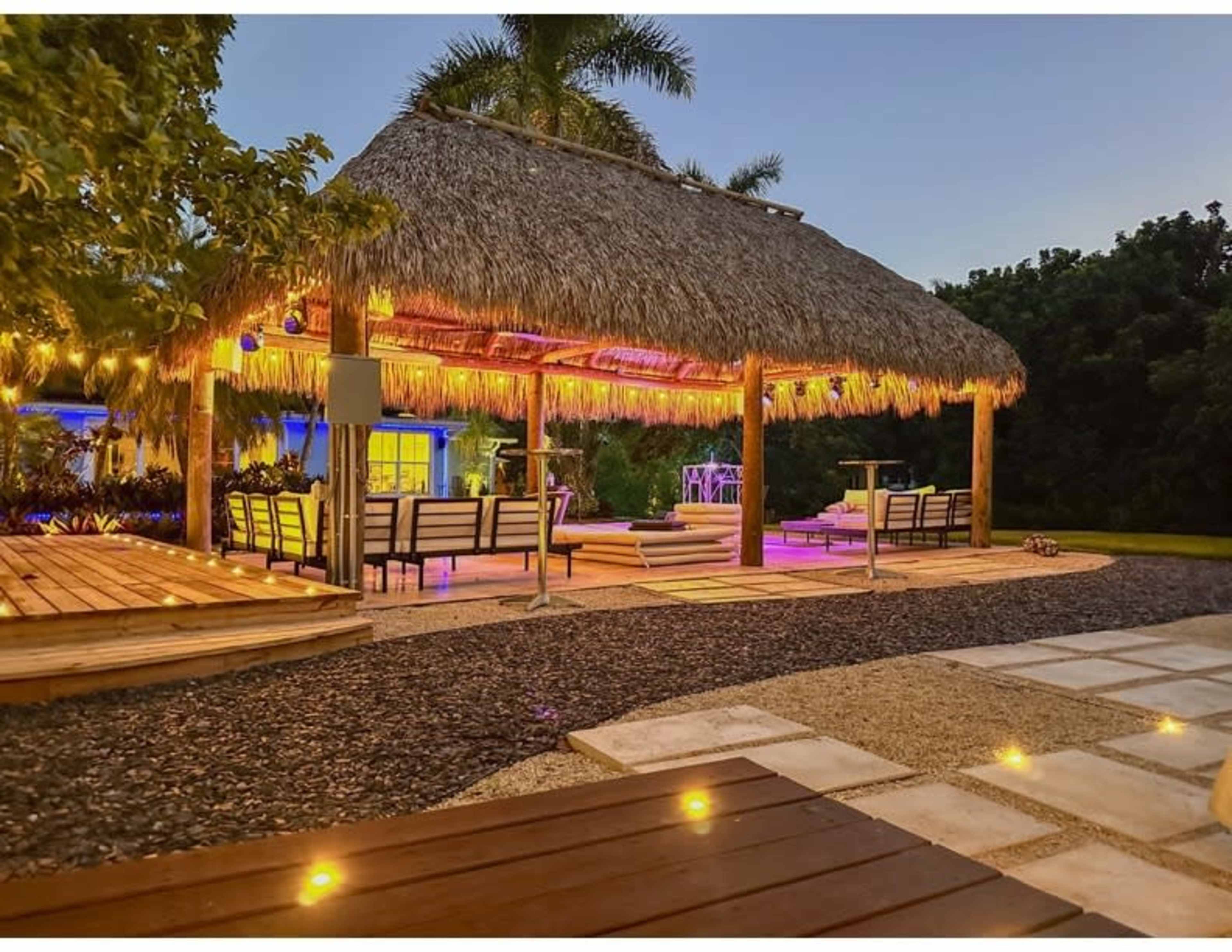 A thatched-roof gazebo with seating and ambient lighting is situated in a landscaped outdoor area.