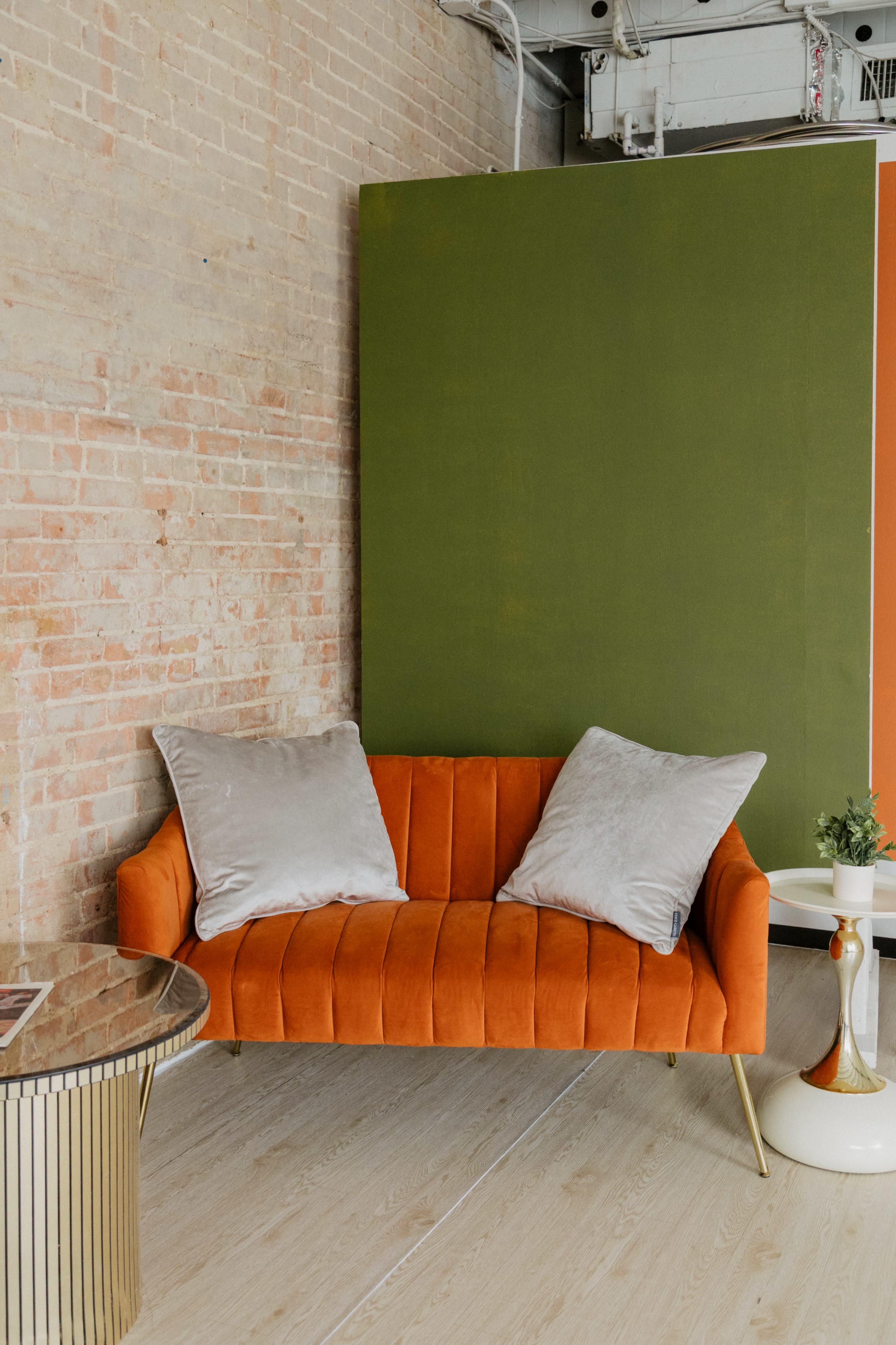The image depicts an orange velvet couch with two gray pillows, situated against a brick wall and a large green backdrop, accompanied by a round glass-topped table.