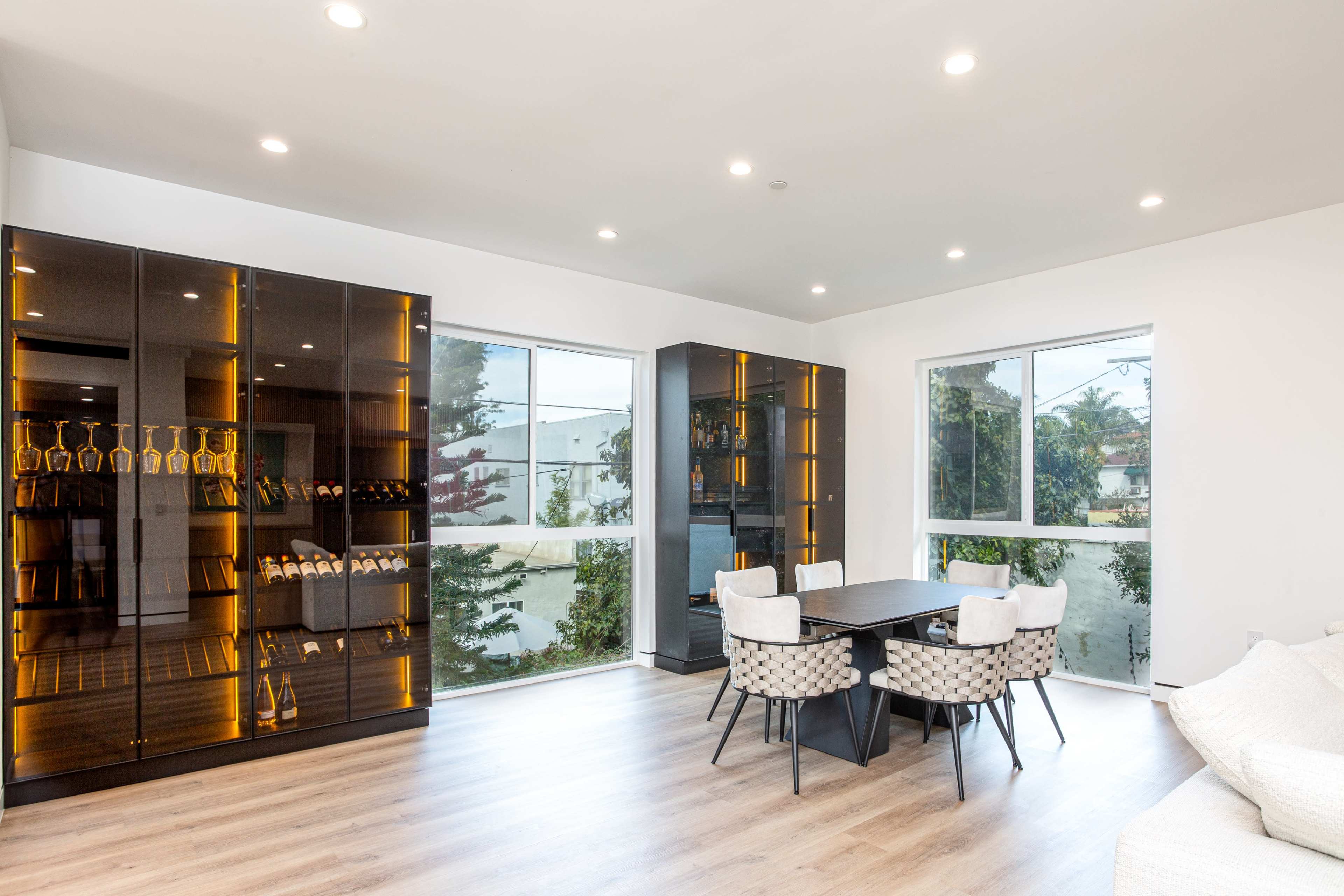 A modern living space with a dining table surrounded by six chairs, glass-front cabinets displaying bottles, and large windows allowing natural light.