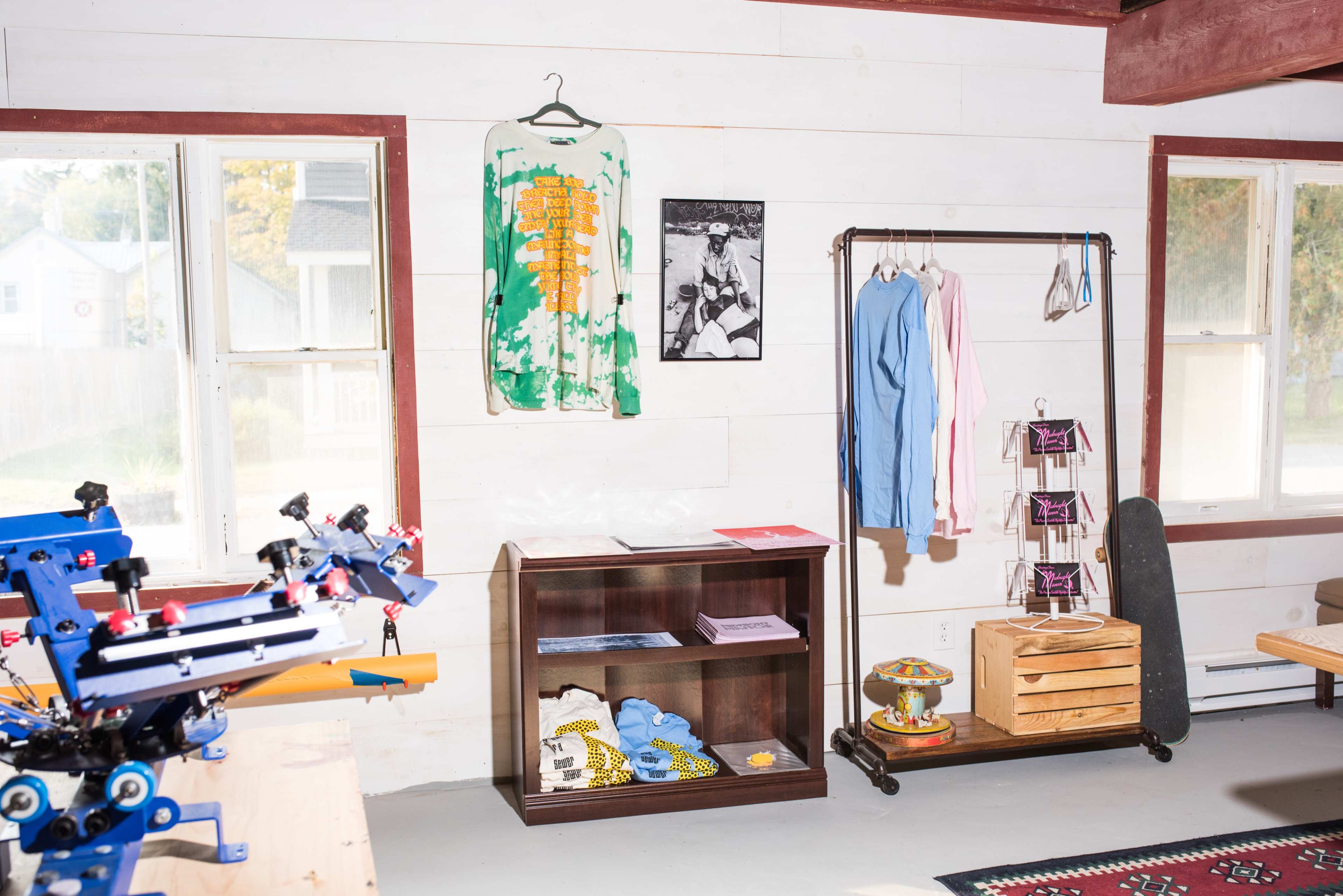 An interior space featuring a clothing display with a variety of garments, a shelf with folded items, and a printing setup on a workbench.