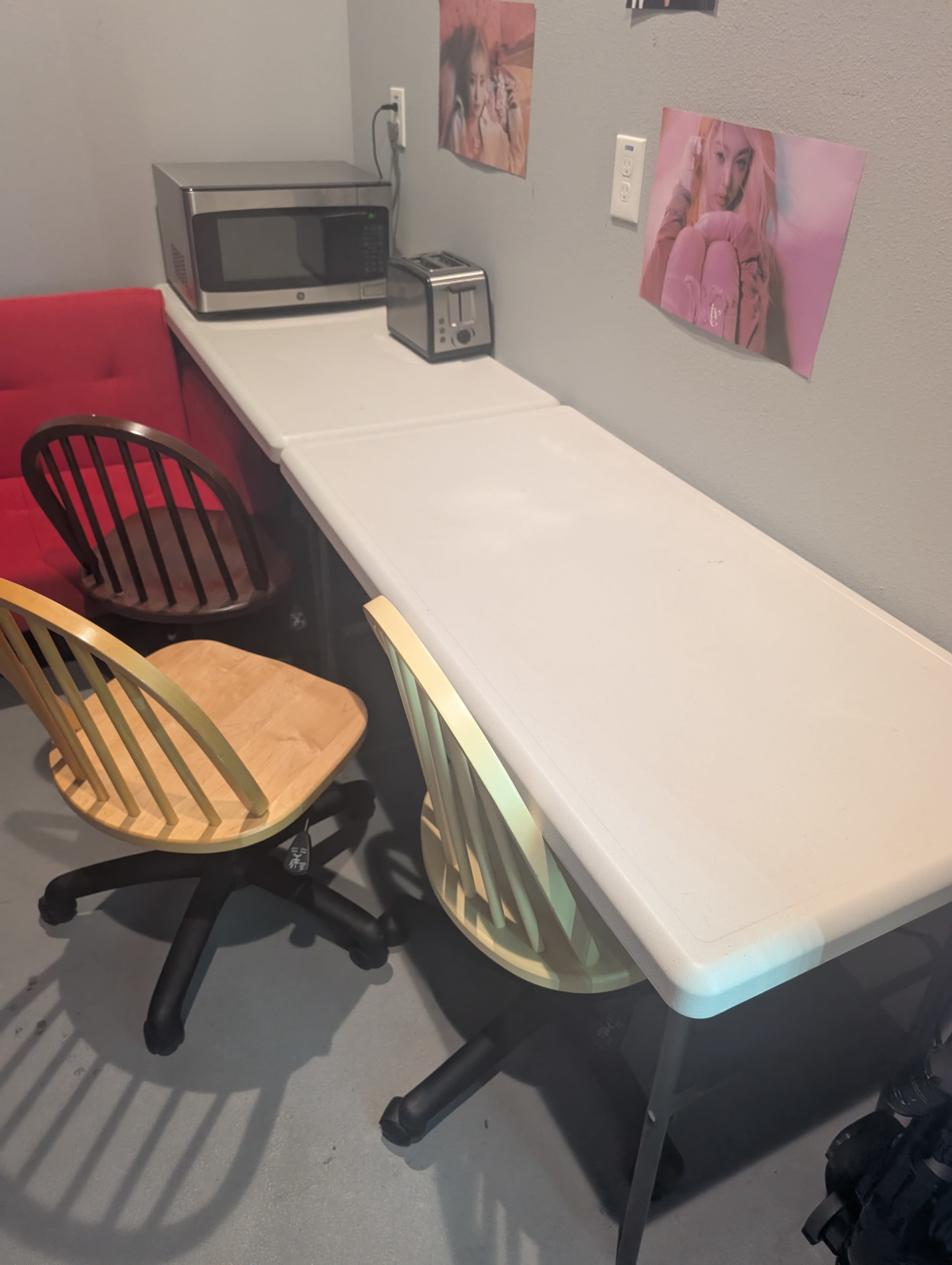 The image shows a small workspace with a long table, two chairs, a toaster, a microwave, and two posters on the wall.