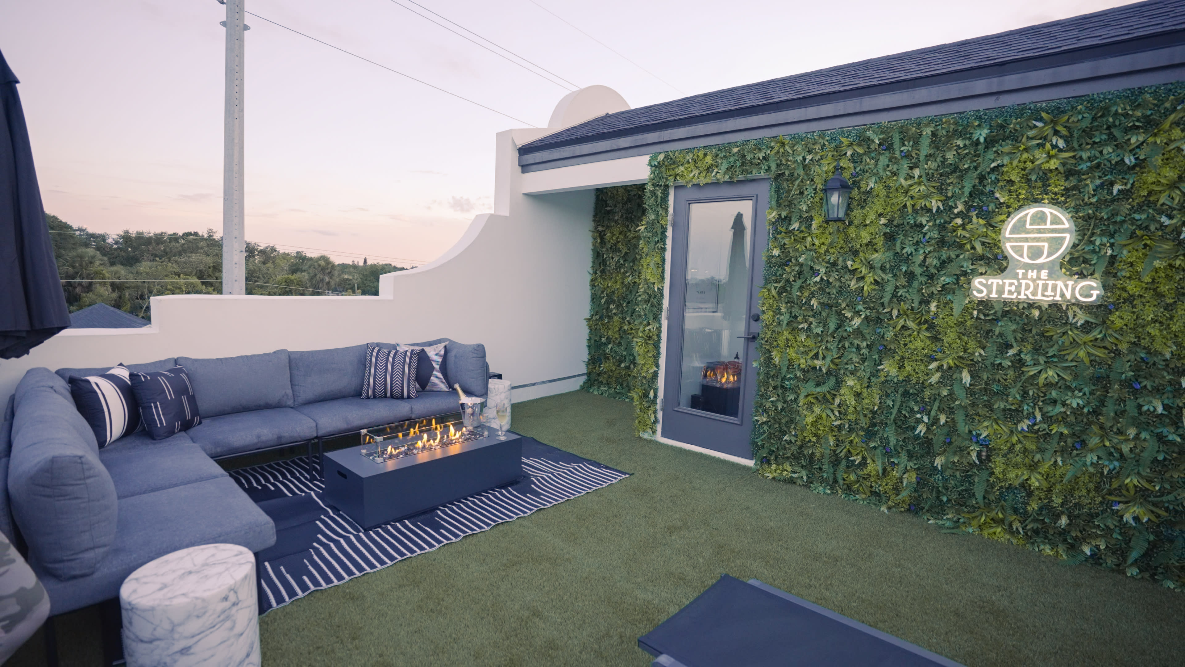 The image shows a rooftop terrace with a blue sectional sofa, a fire table, and a wall covered in greenery, along with a door and a logo on the wall.