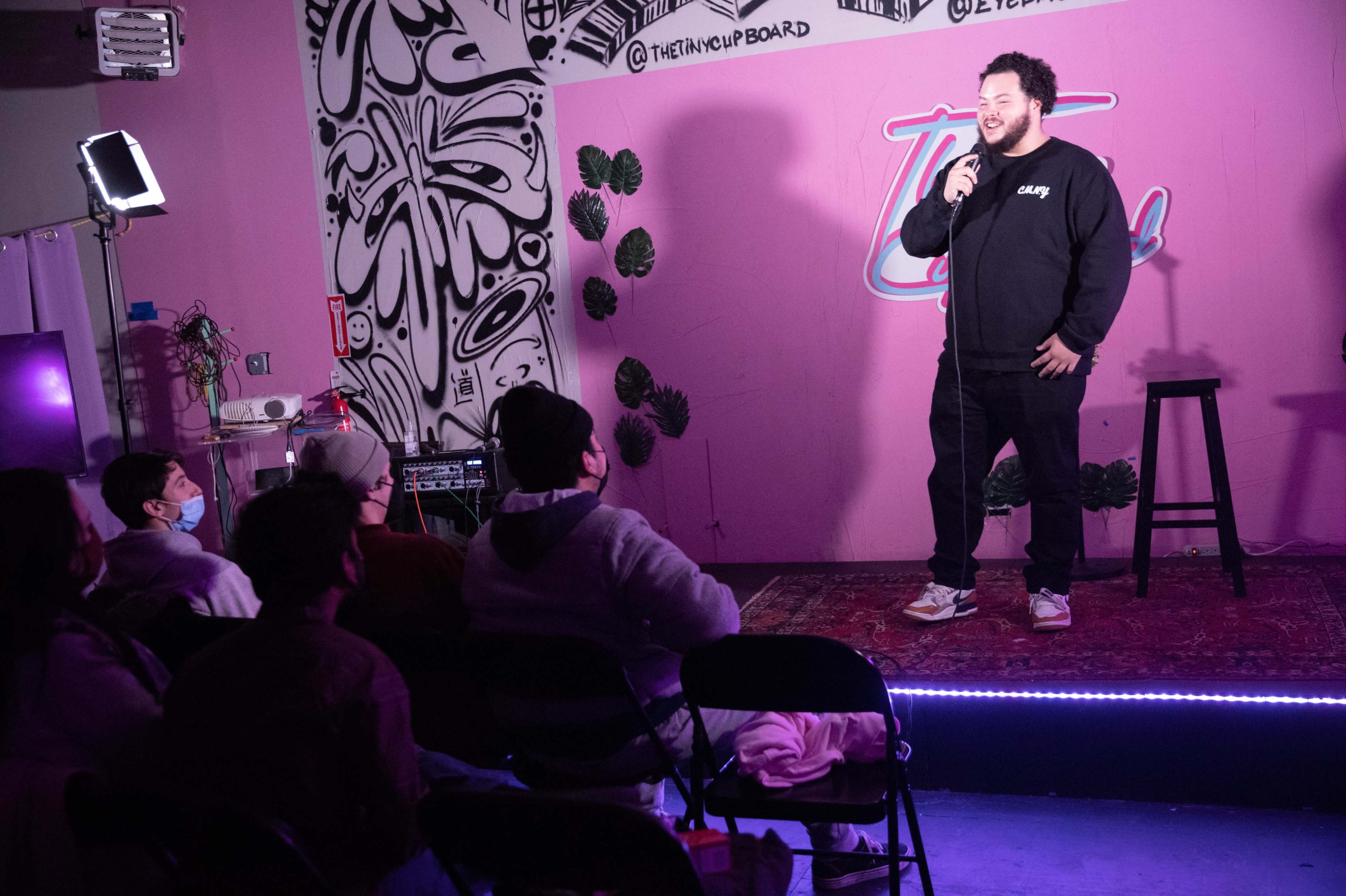 A comedian performs on stage in a pink-walled venue, while a small audience watches from seated chairs.