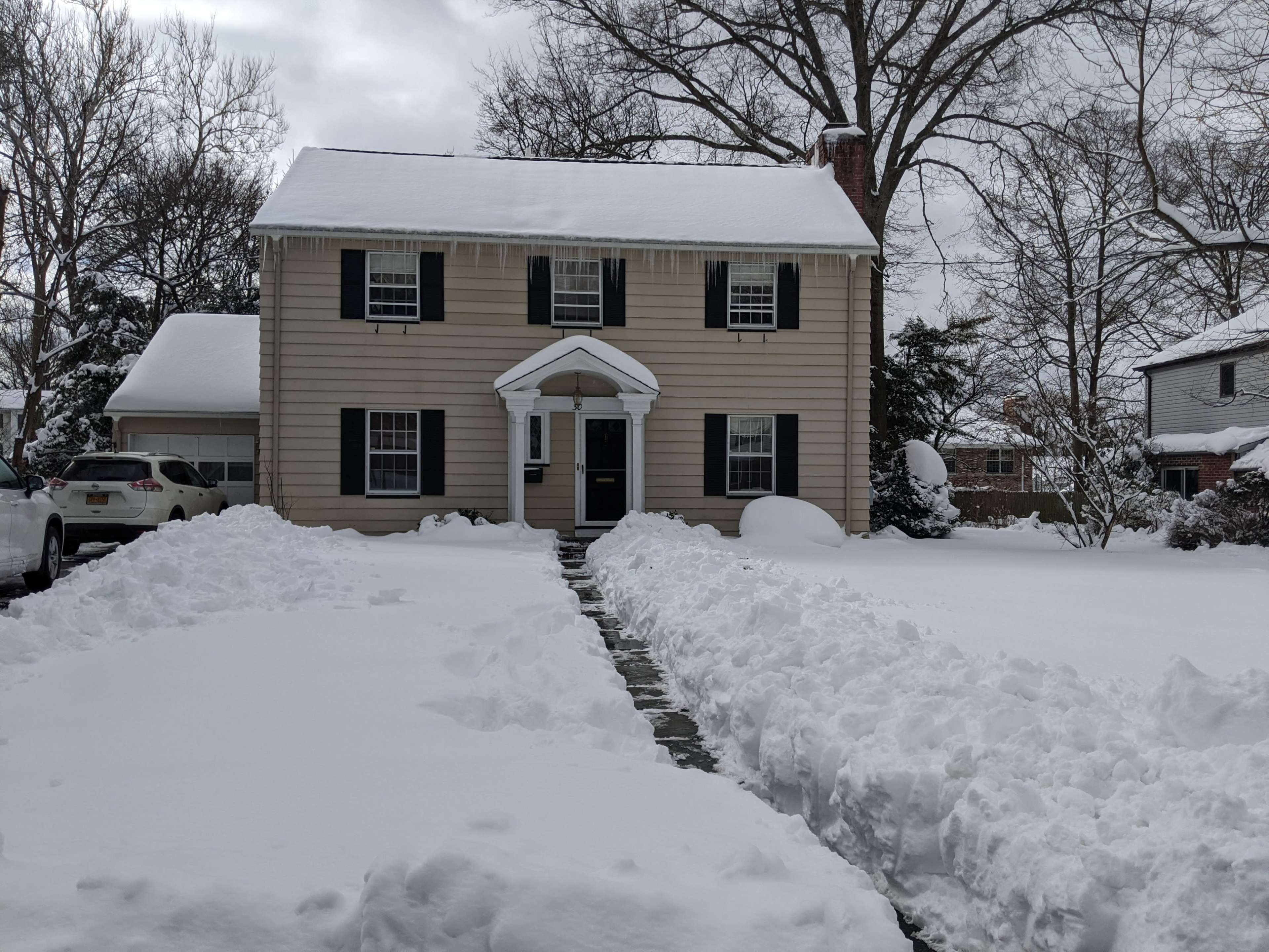 A two-story house with black shutters and a front walkway is surrounded by a thick layer of freshly fallen snow.
