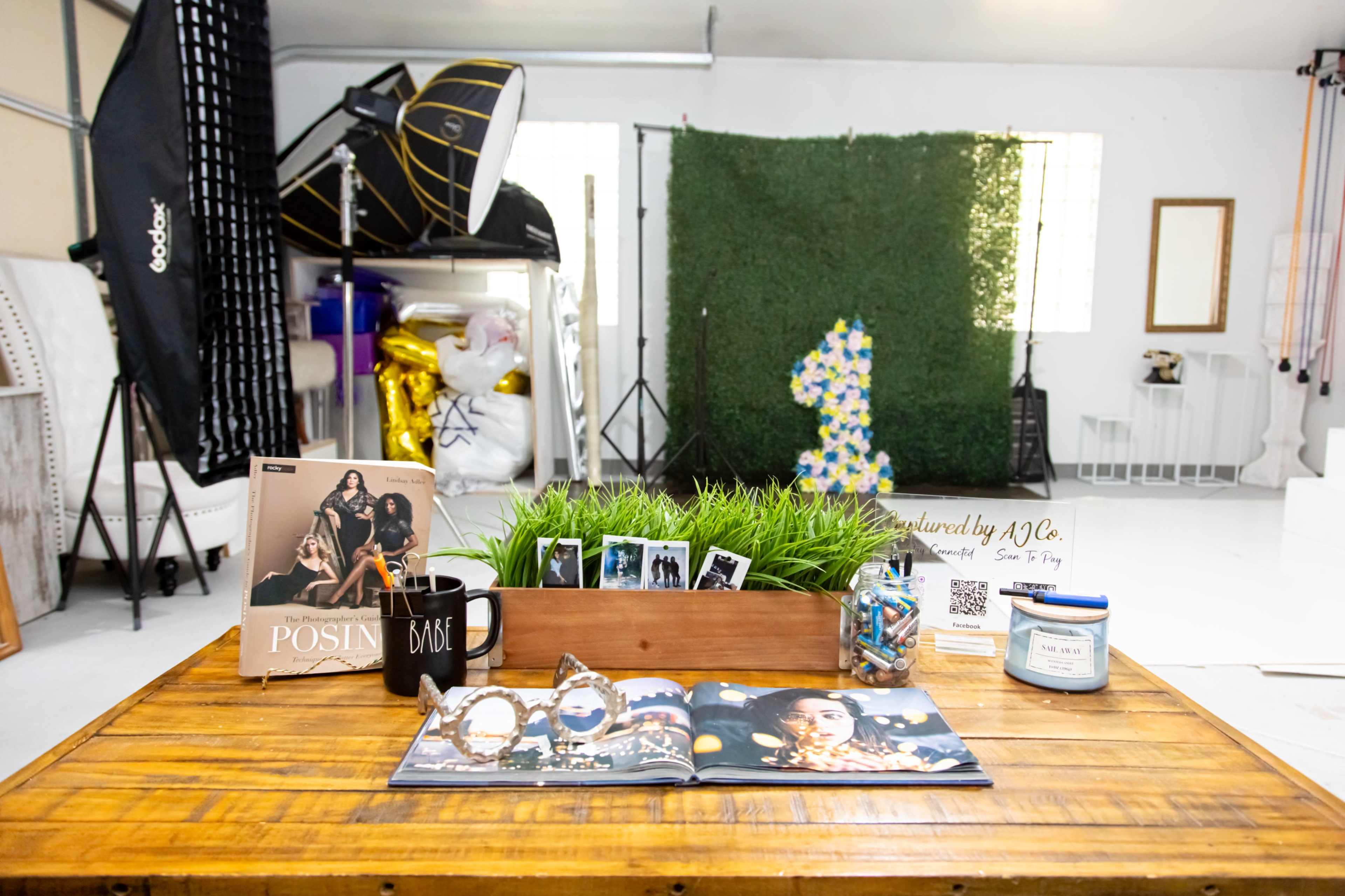 The image shows a studio setup featuring a wooden table with a magazine, photo prints, a mug, and a notebook, surrounded by photography equipment and a decorative backdrop.