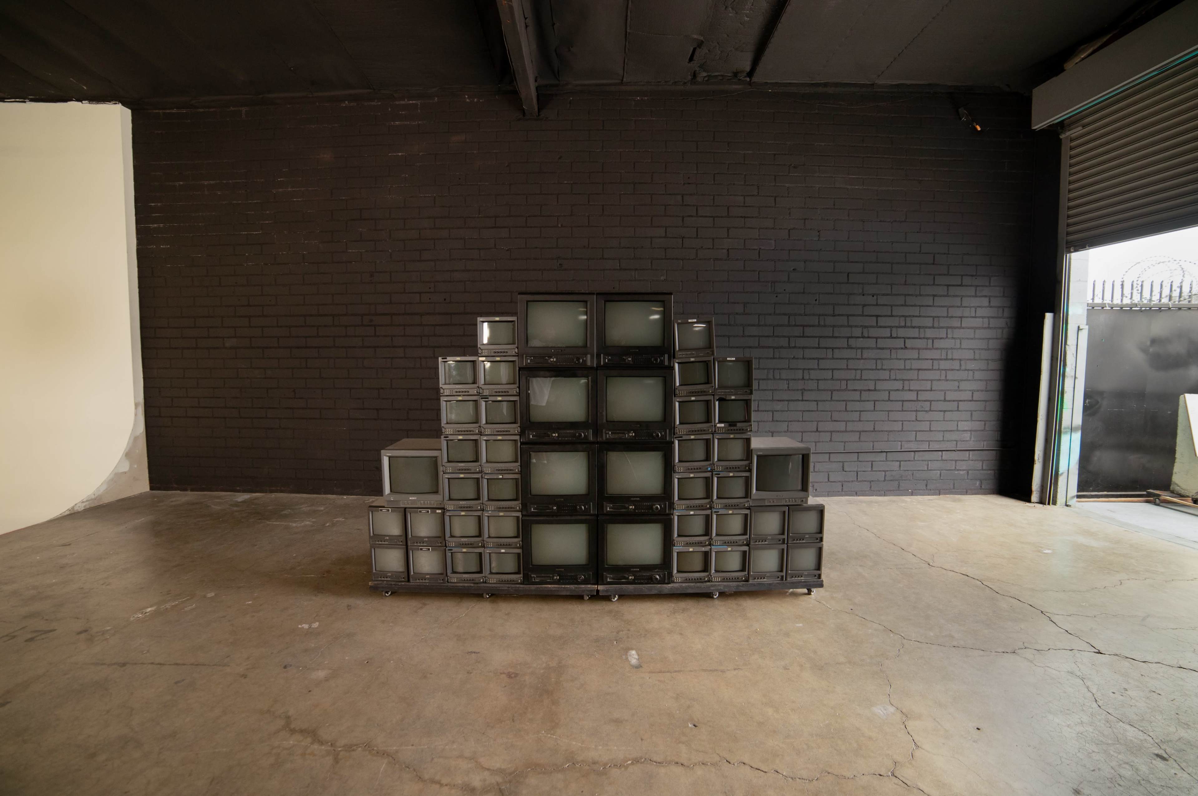A pyramid-shaped structure made of stacked old televisions is positioned in a spacious, industrial-style room with a black brick wall.