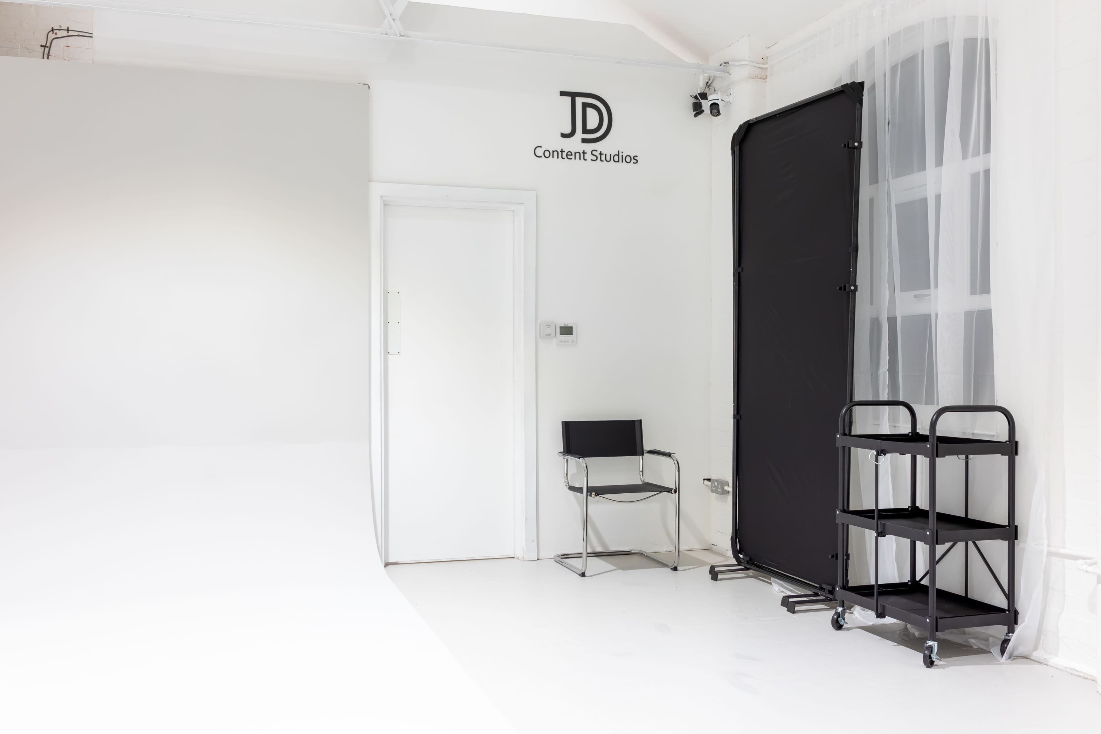The image shows a minimalist studio space with a simple chair, a portable cart, and a backdrop, featuring the logo "JD Content Studios" on the wall.