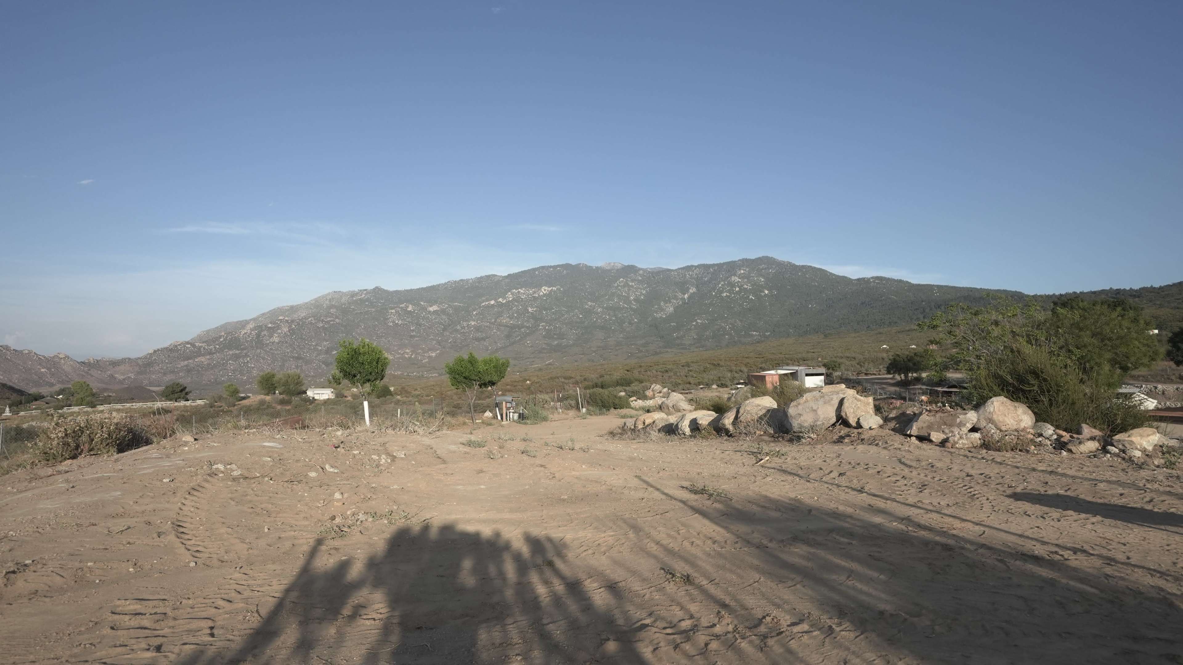 The image shows a sandy landscape with scattered rocks, shrubbery, and a mountain in the background under a clear blue sky.