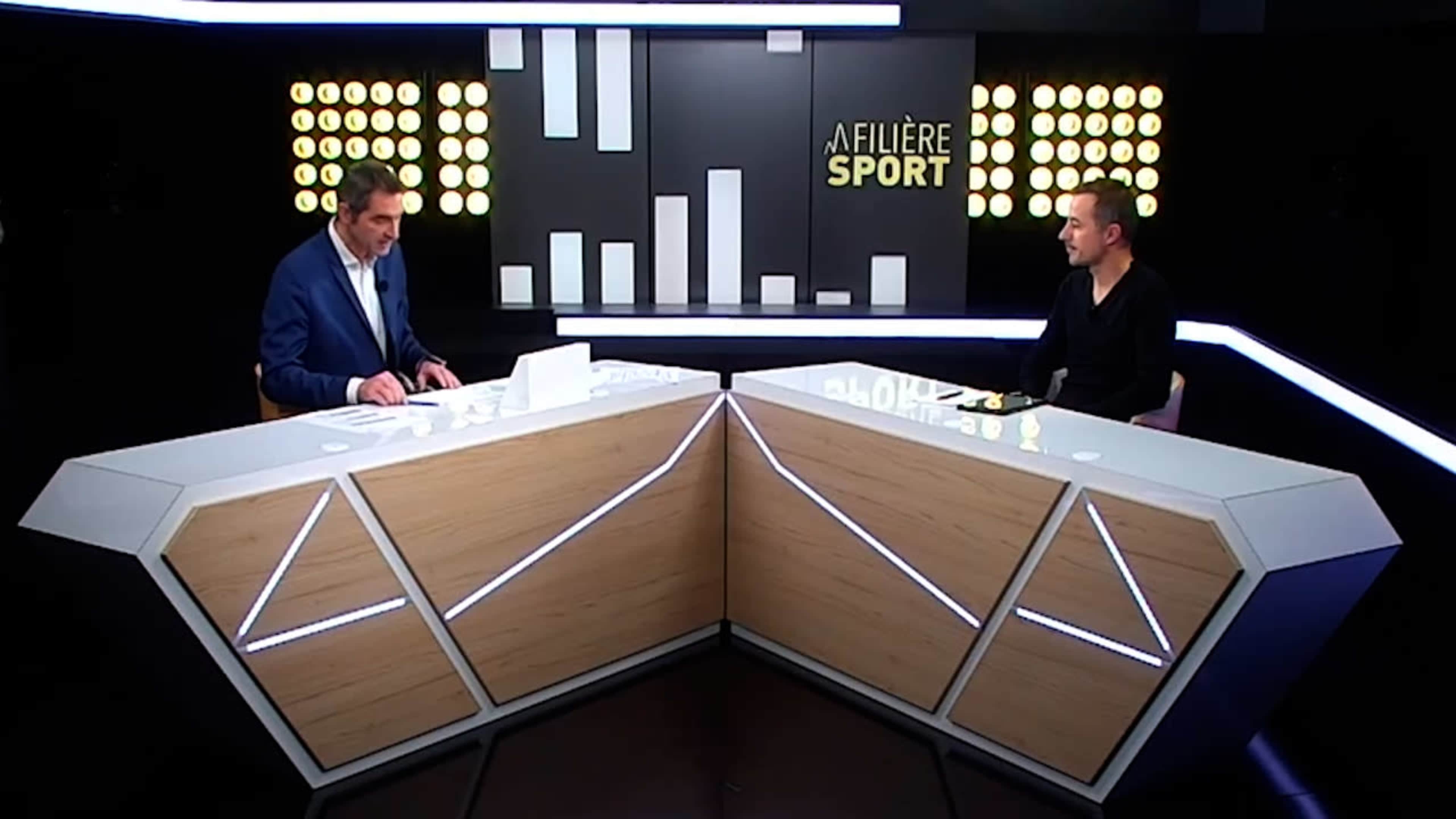 Two men are seated at a modern news desk with a backdrop displaying the title "Filière Sport" and illuminated circular lights.