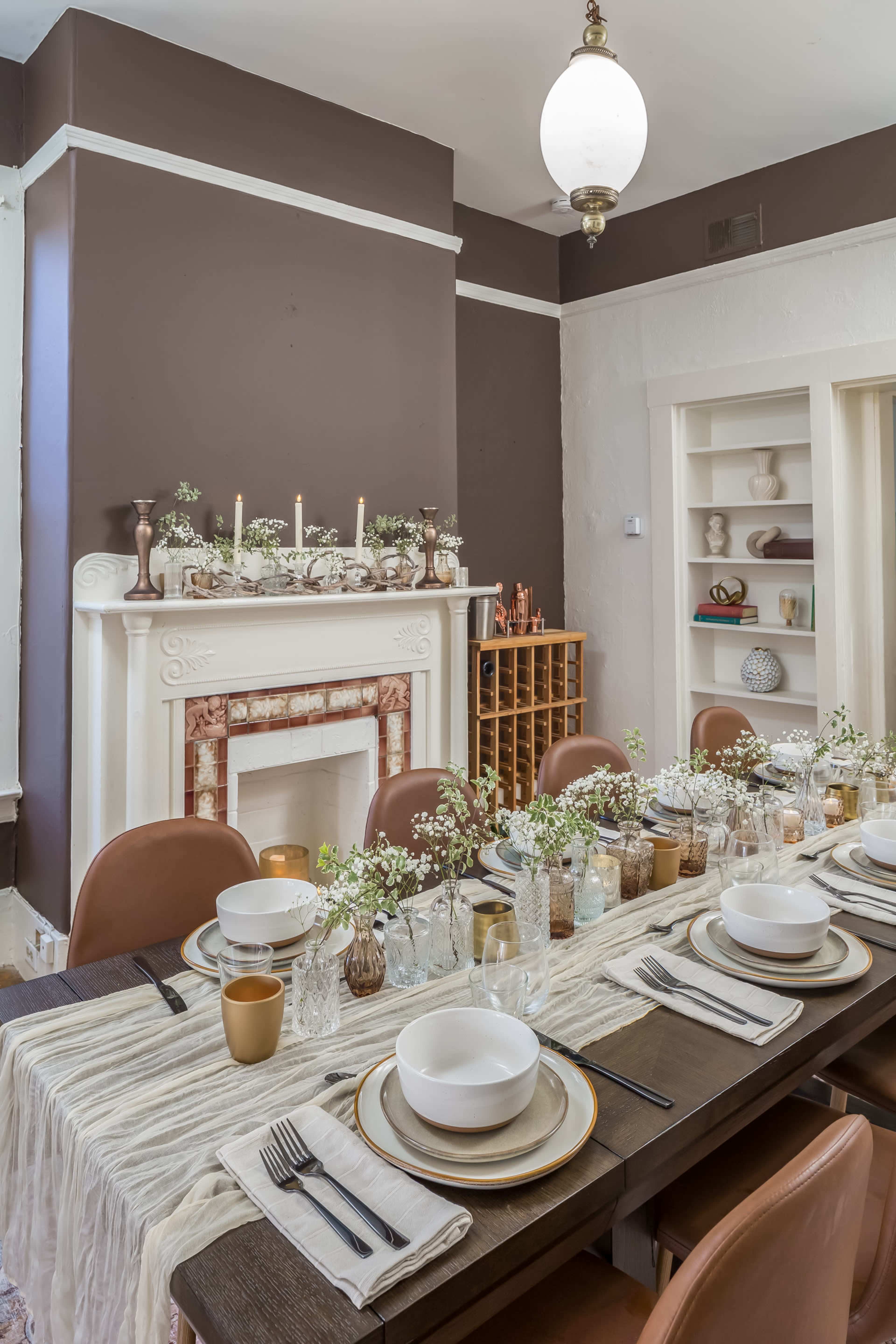 A dining room features a long wooden table set with dishware and decorative flowers, complemented by a fireplace adorned with candles and a shelf displaying various objects.