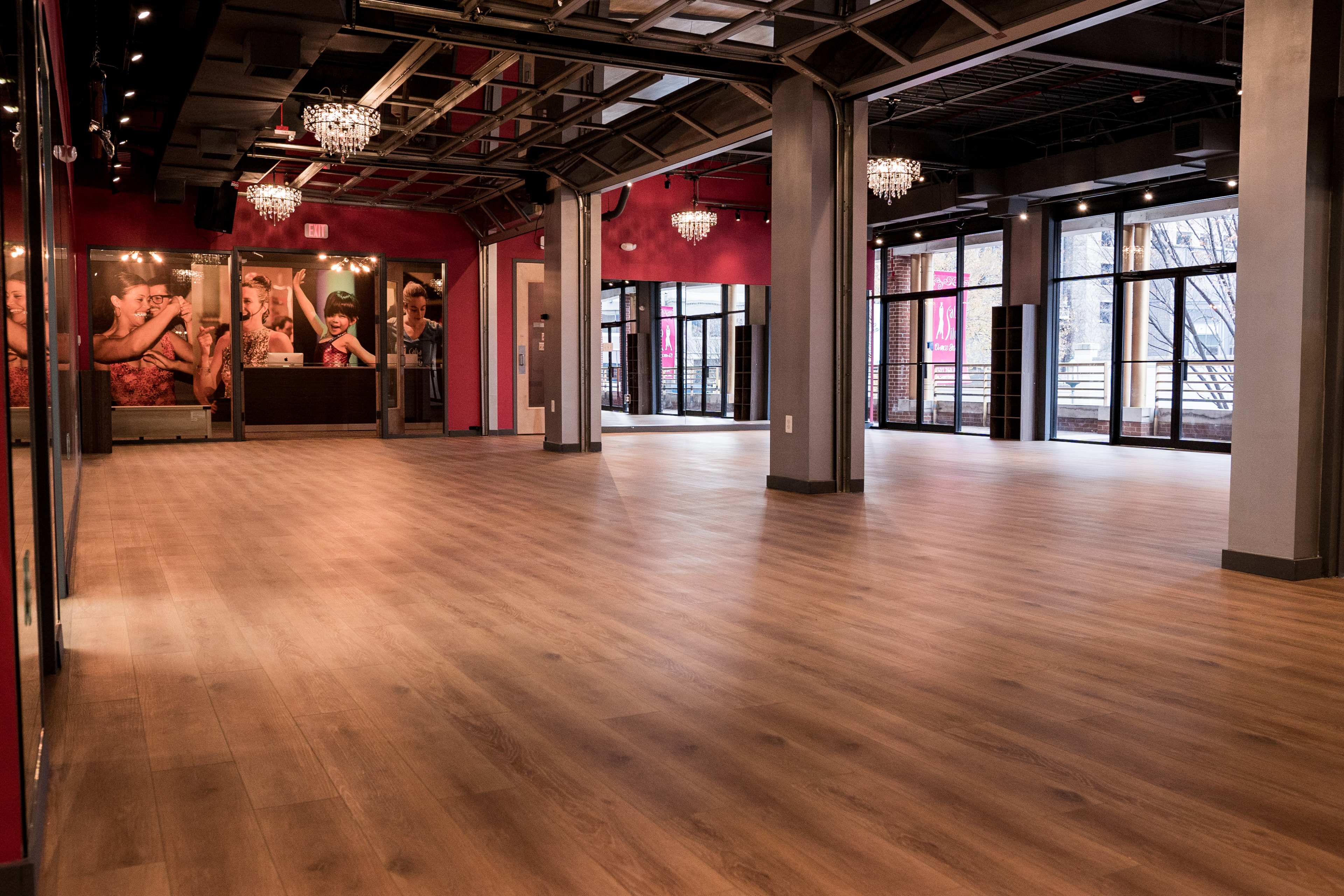 A spacious interior room with a wooden floor, high ceilings, and large windows, featuring chandeliers and vibrant red walls.