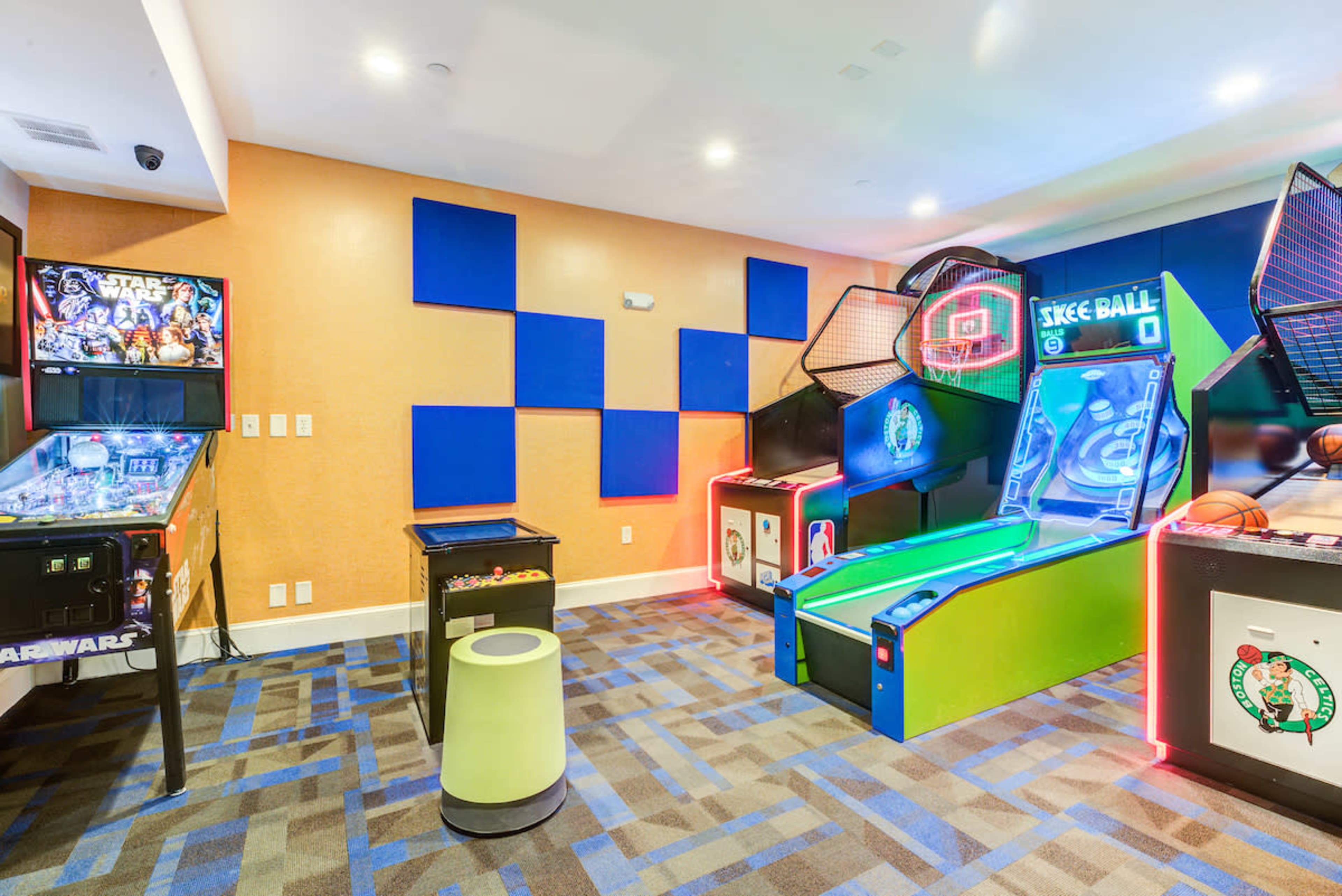 A game room featuring a pinball machine, a basketball arcade game, and a skeeball machine, with colorful wall panels and carpet.