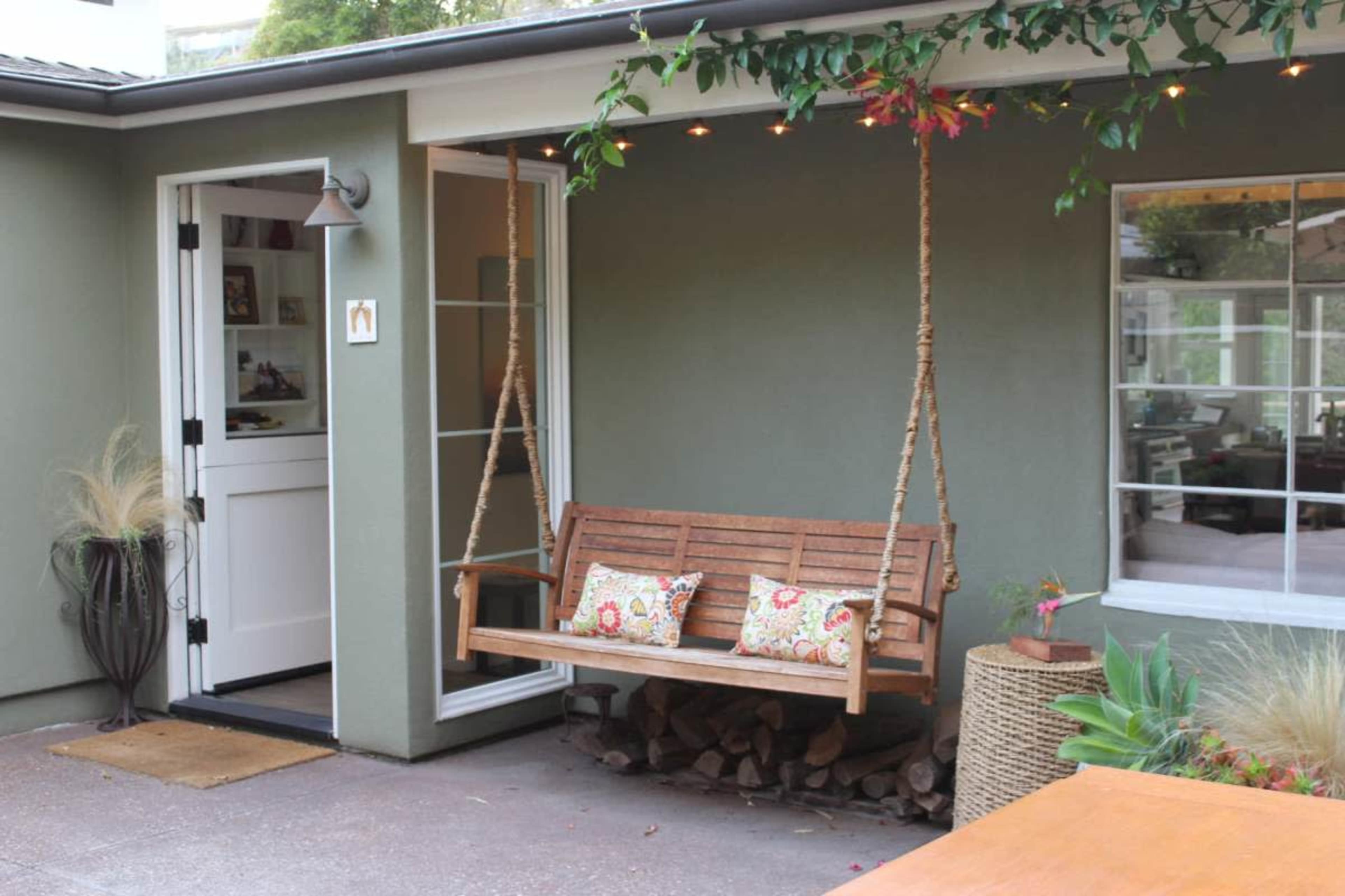 A wooden swing bench with decorative cushions is suspended from a porch awning next to a door and plants.