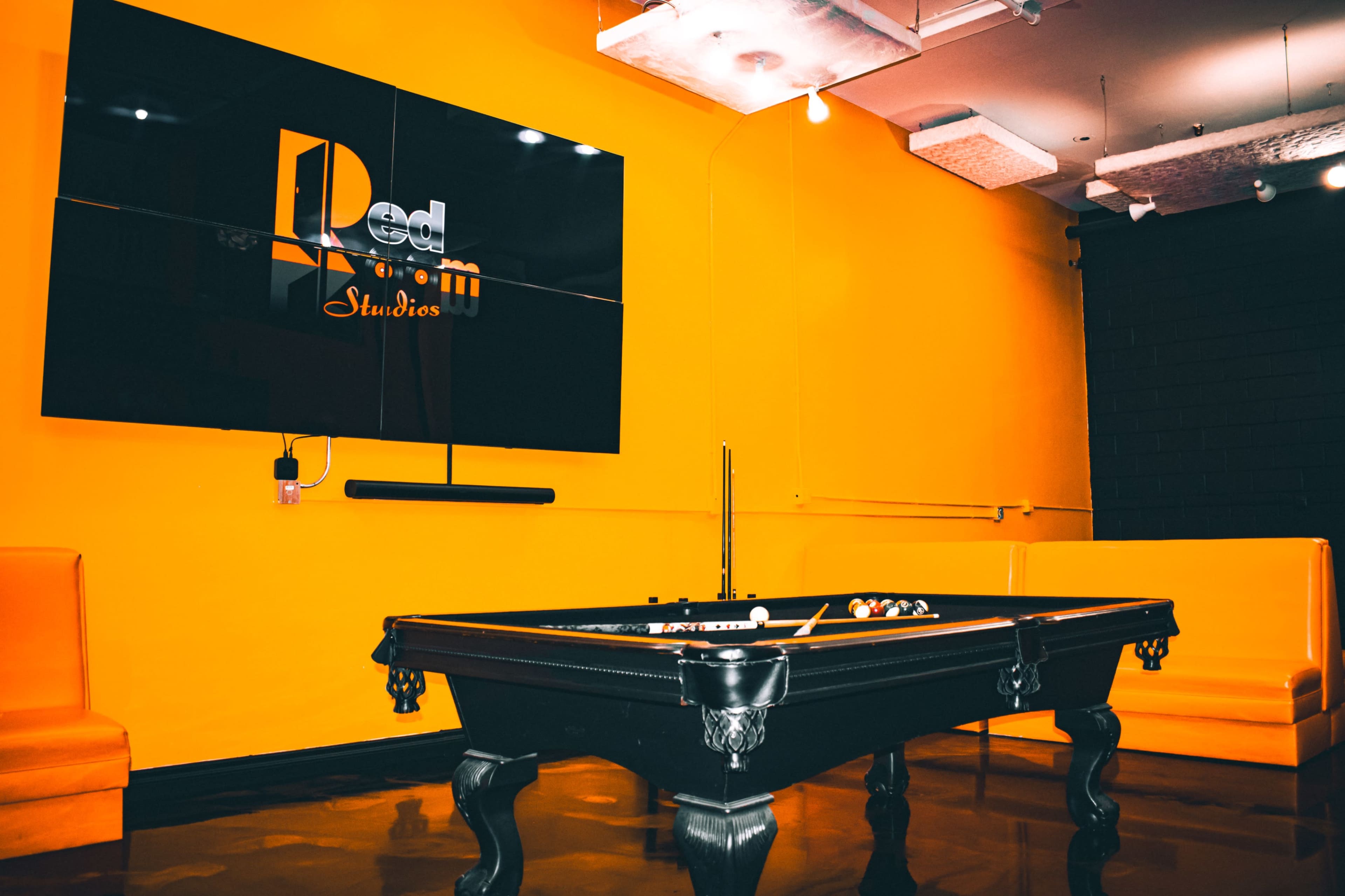 A black pool table is centered in a room with bright orange walls, featuring a large screen displaying the logo for "Red Studios."