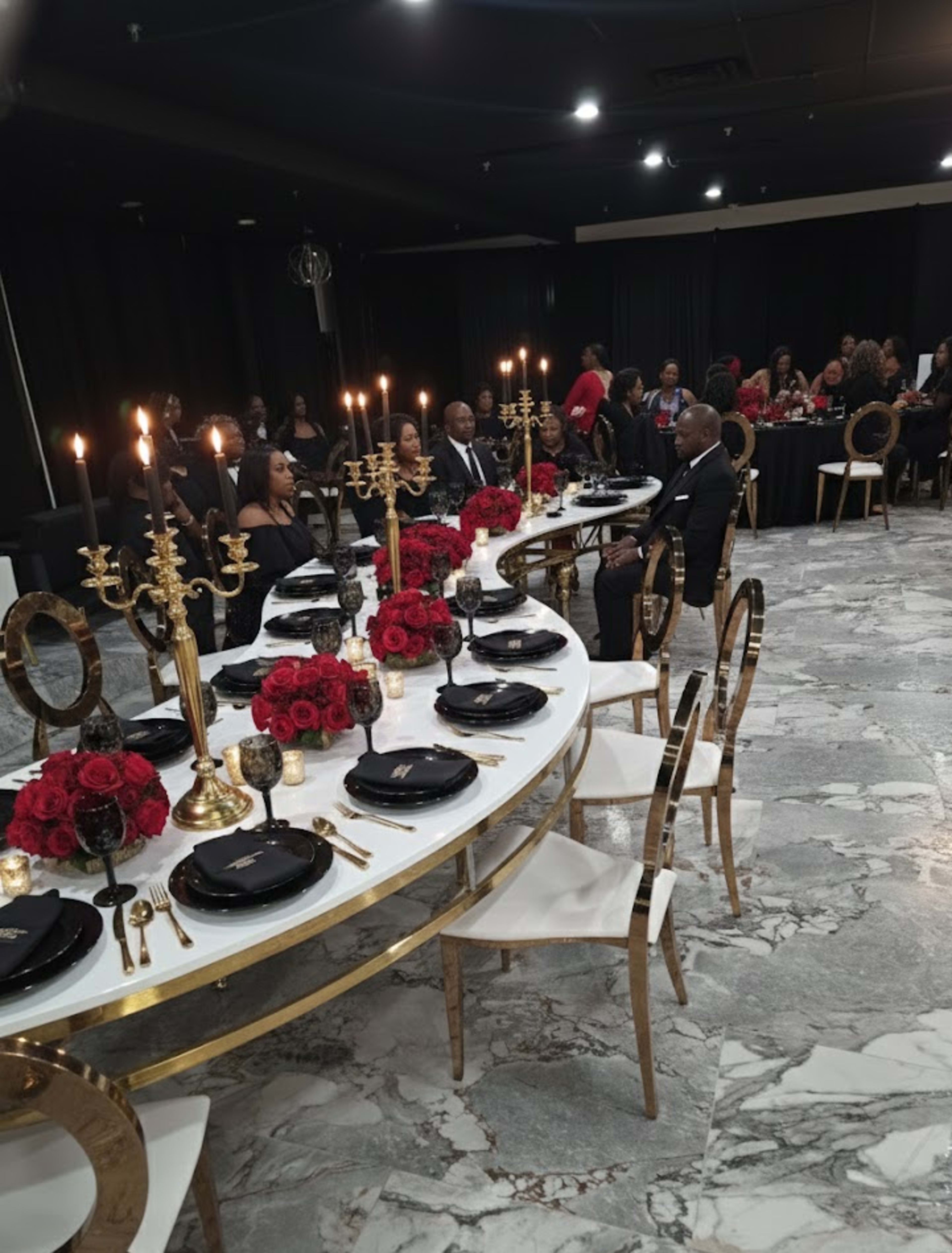 A long, elegantly set dinner table with gold accents and red floral centerpieces is occupied by guests dressed in formal attire at a sophisticated event.