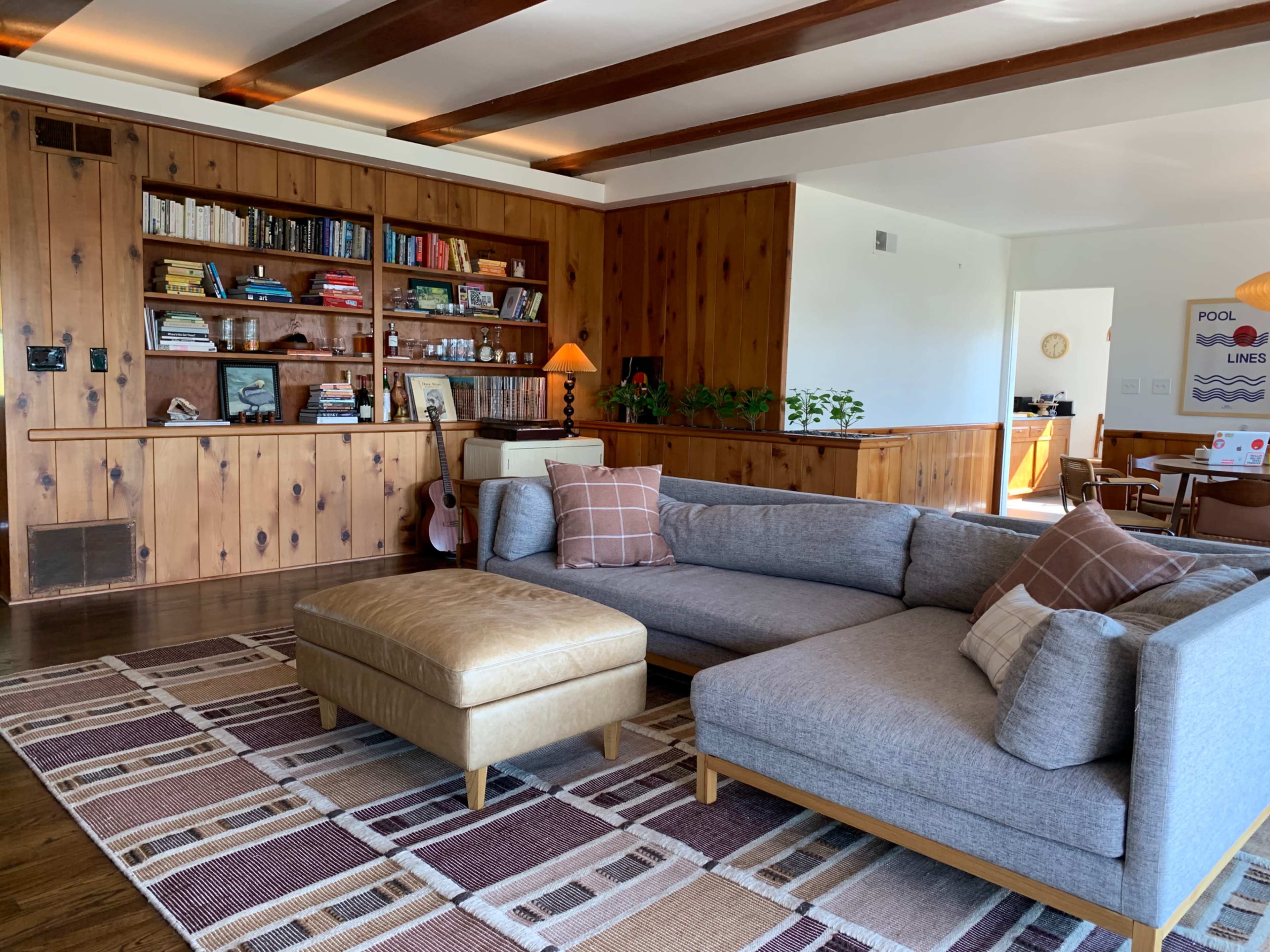 A modern living room features a large gray sectional sofa and a leather ottoman, with wooden paneling and built-in shelves filled with books in the background.