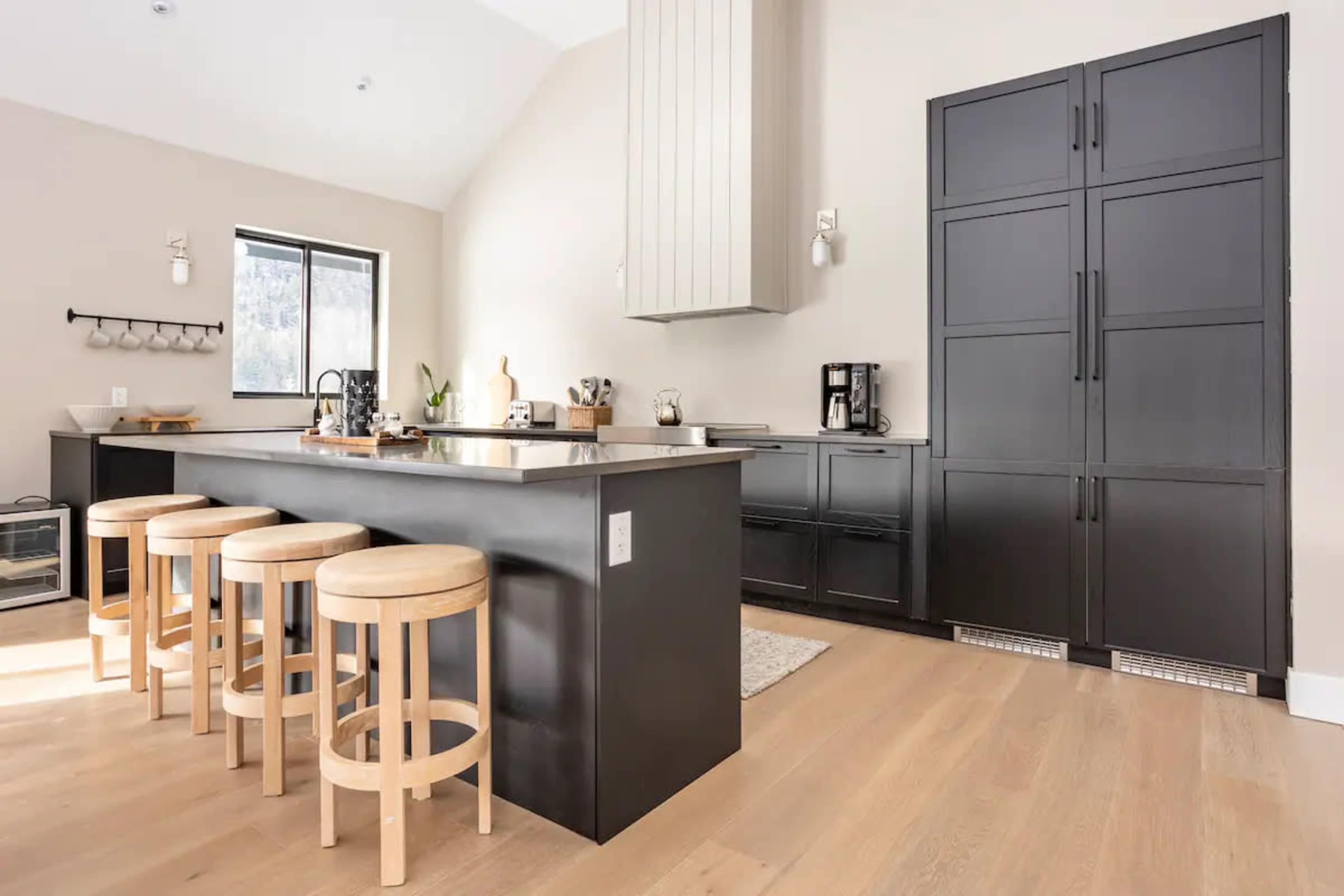 A modern kitchen features a large island with four wooden stools, sleek black cabinetry, and stainless steel appliances.