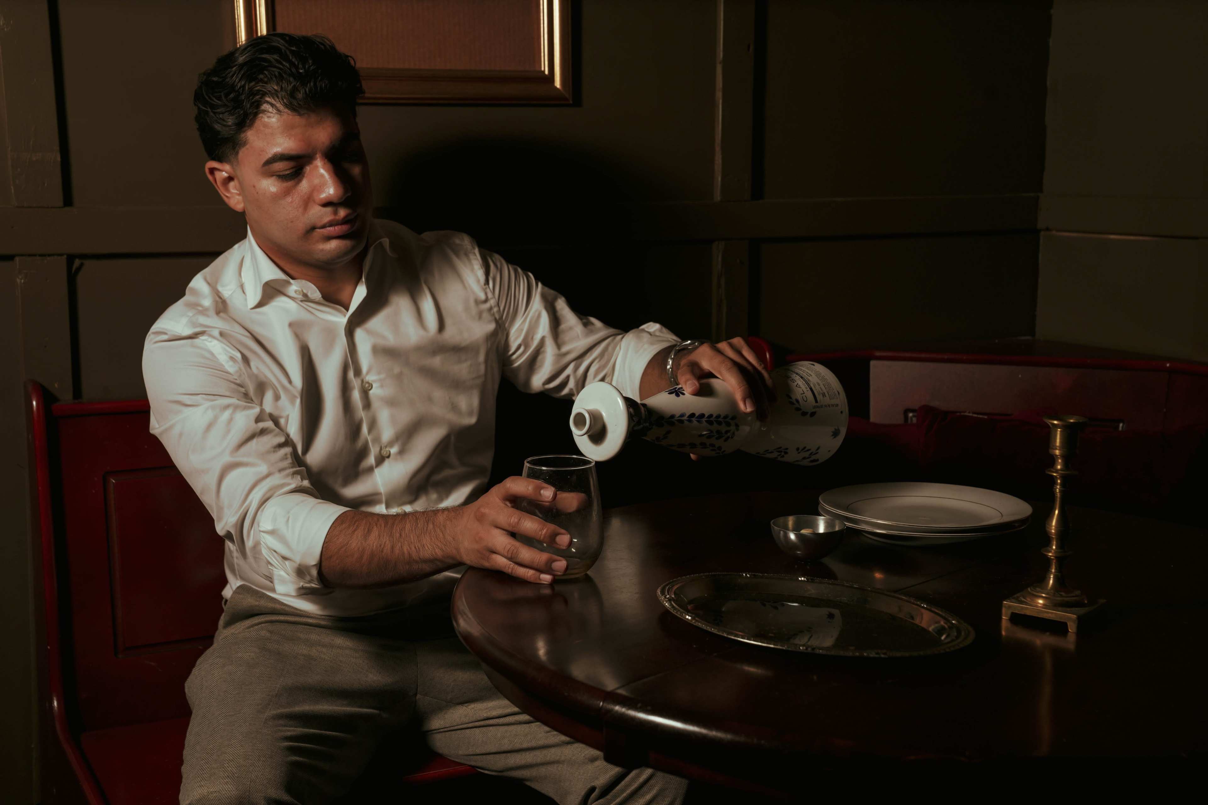 A man in a white shirt pours a drink from a bottle into a glass on a wooden table.