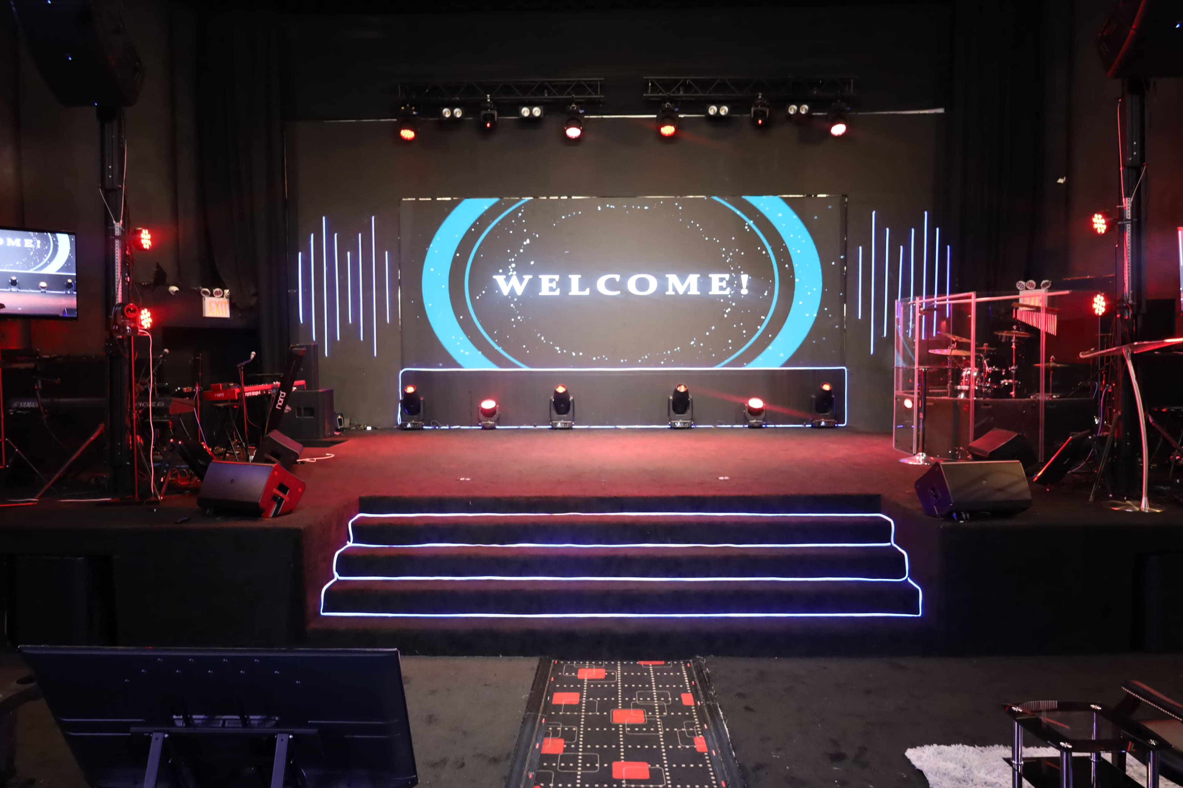 The image shows a stage set up for an event, featuring a large screen with the word "WELCOME," colorful lighting, and musical equipment.