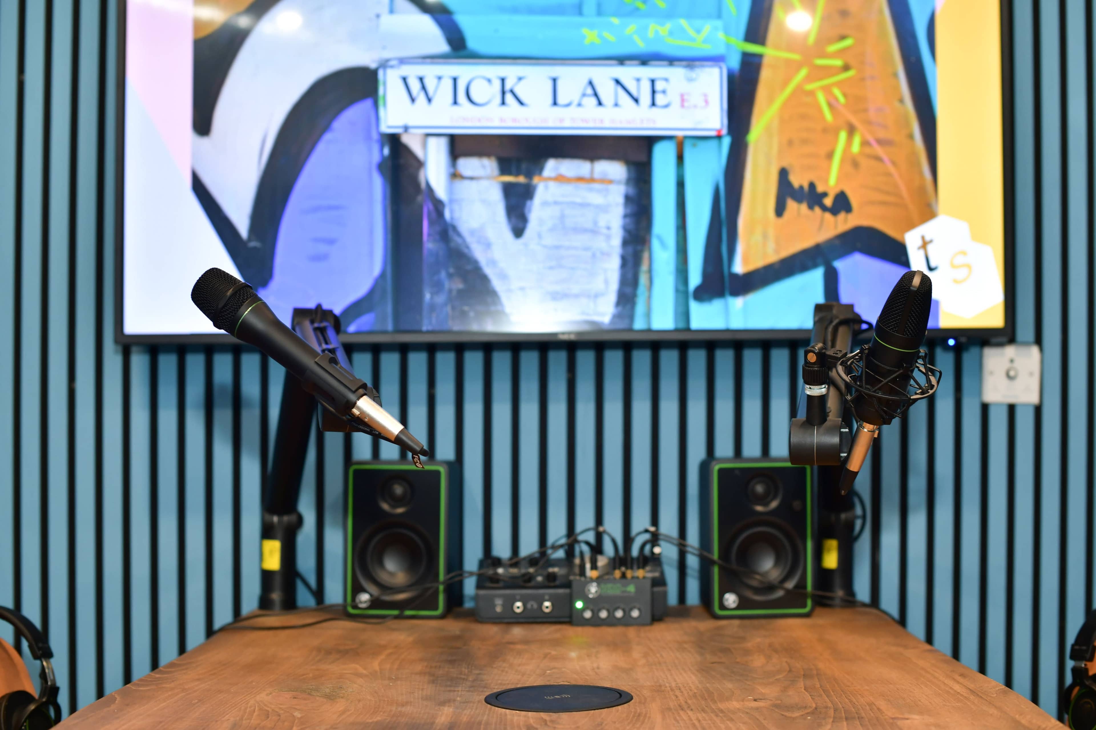 A podcast setup featuring two microphones on adjustable stands positioned over a wooden table, with audio equipment and a colorful mural in the background.