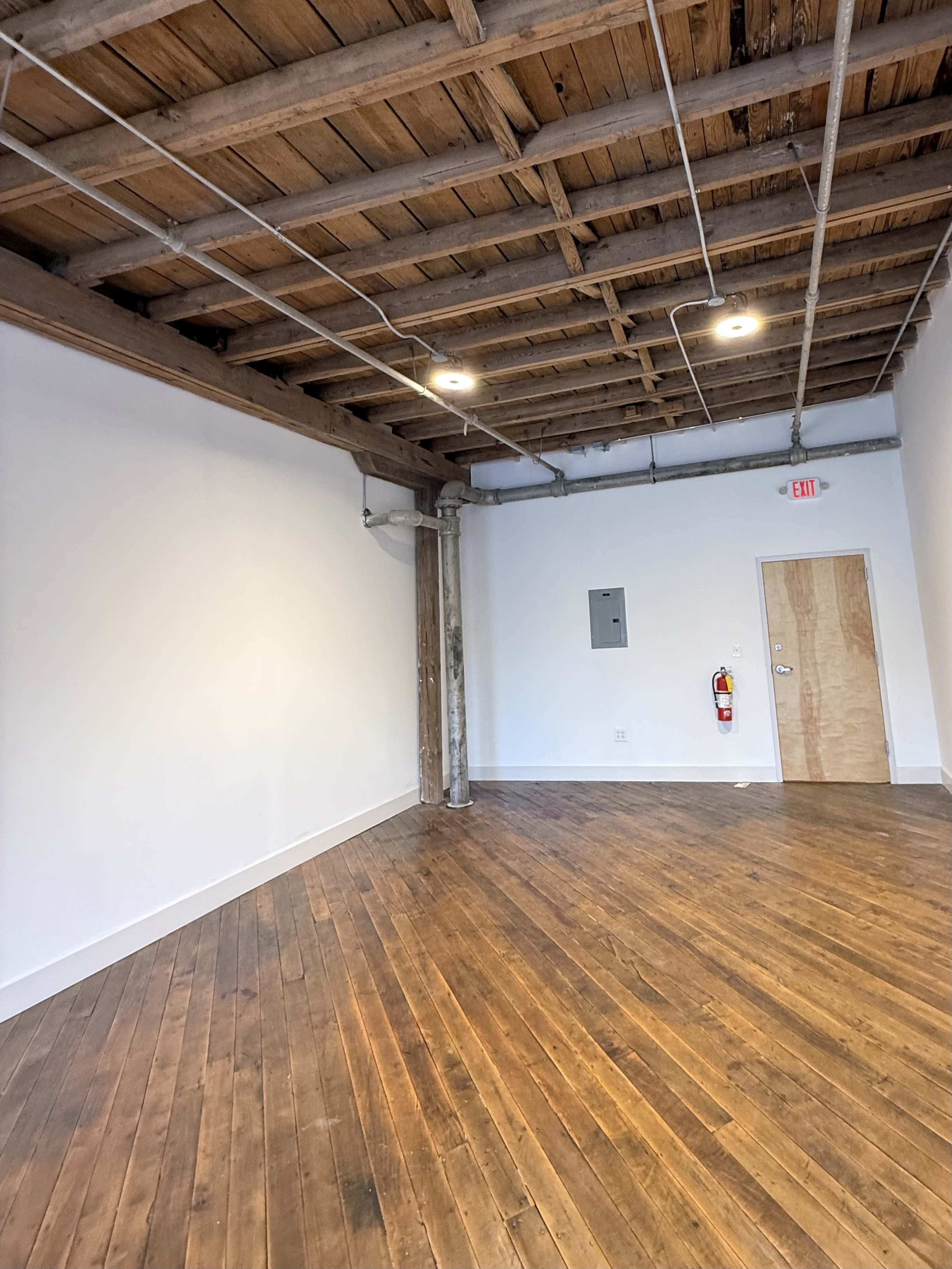 The image shows an empty room with wooden flooring, exposed beams, and a simple design, featuring a door and a wall-mounted electrical panel.
