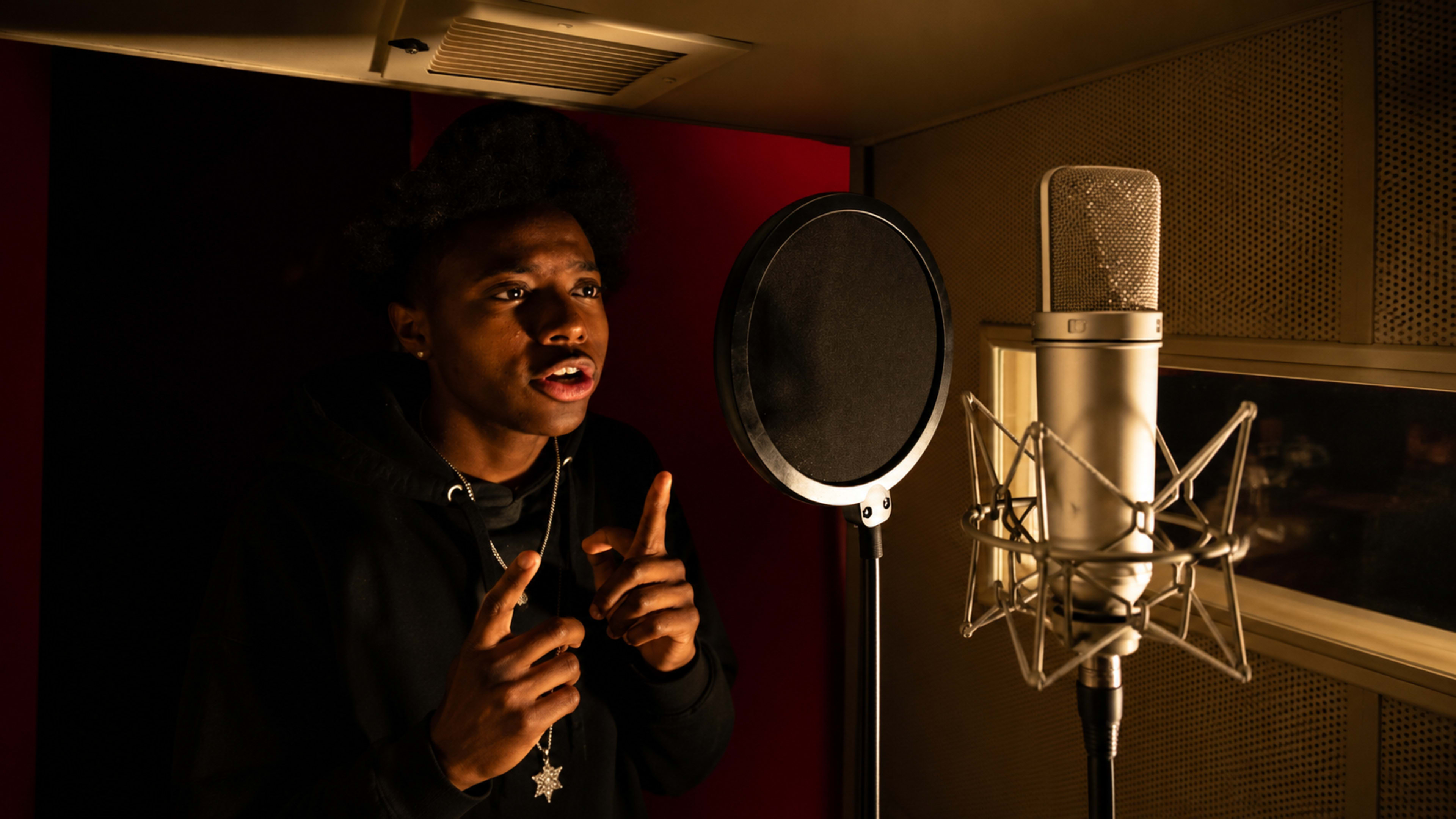 A young man is recording in a sound booth, standing near a microphone with a focused expression.