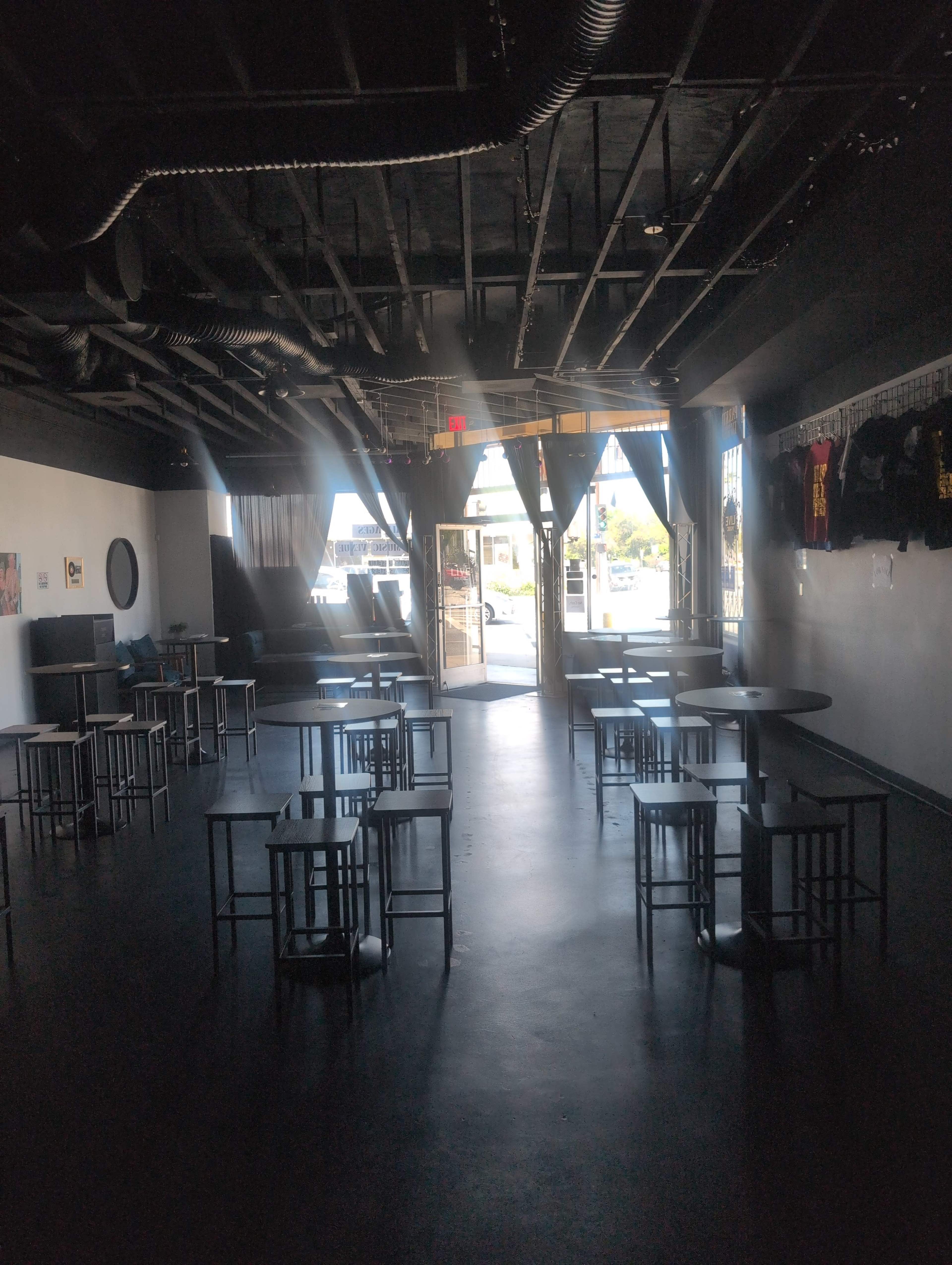 The image shows an empty room with several tall tables and chairs, leading to an open doorway with curtains.