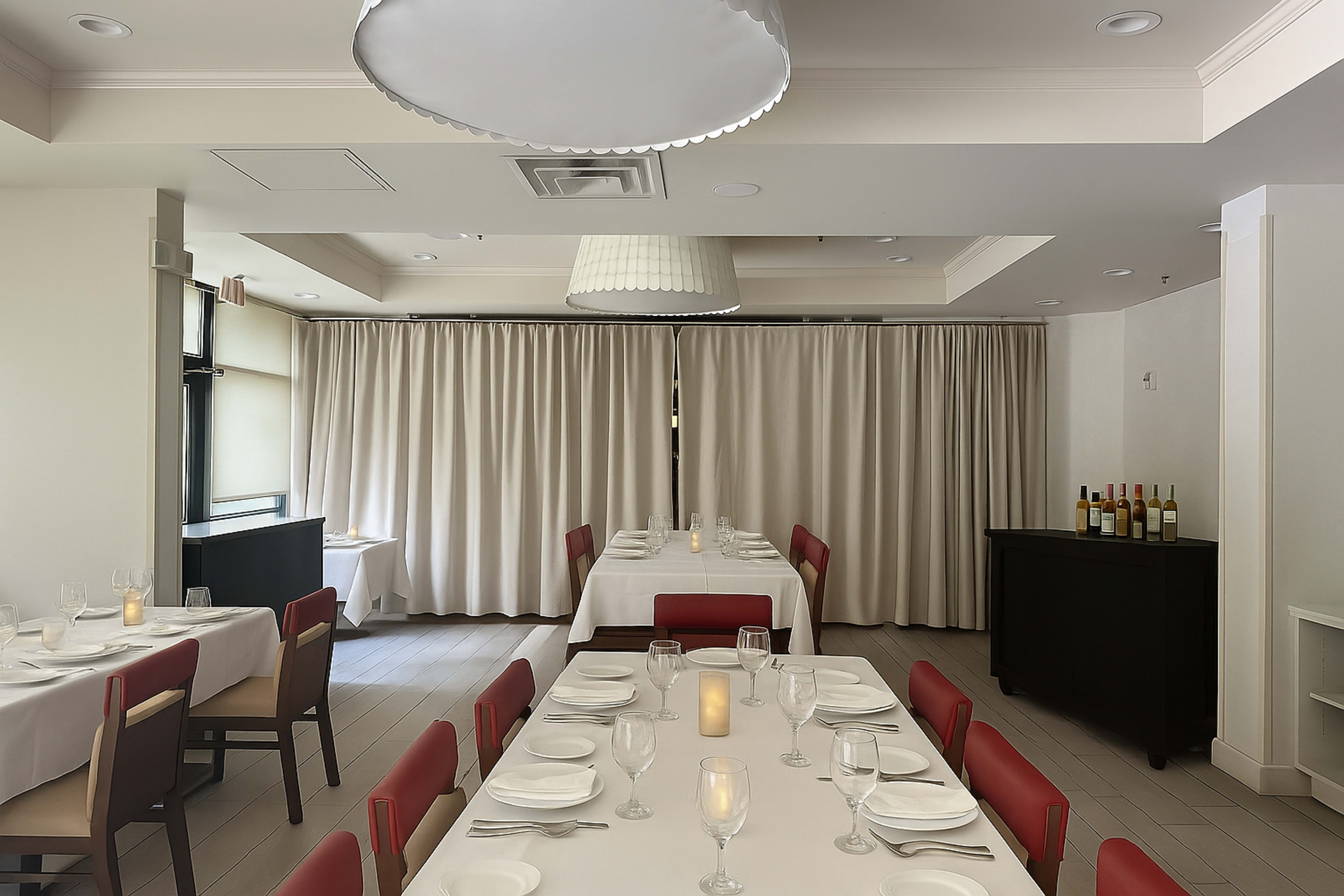 The image shows a modern restaurant interior with white tablecloths, red chairs, and a curtained window, featuring a candlelit table setting and a wine display in the corner.