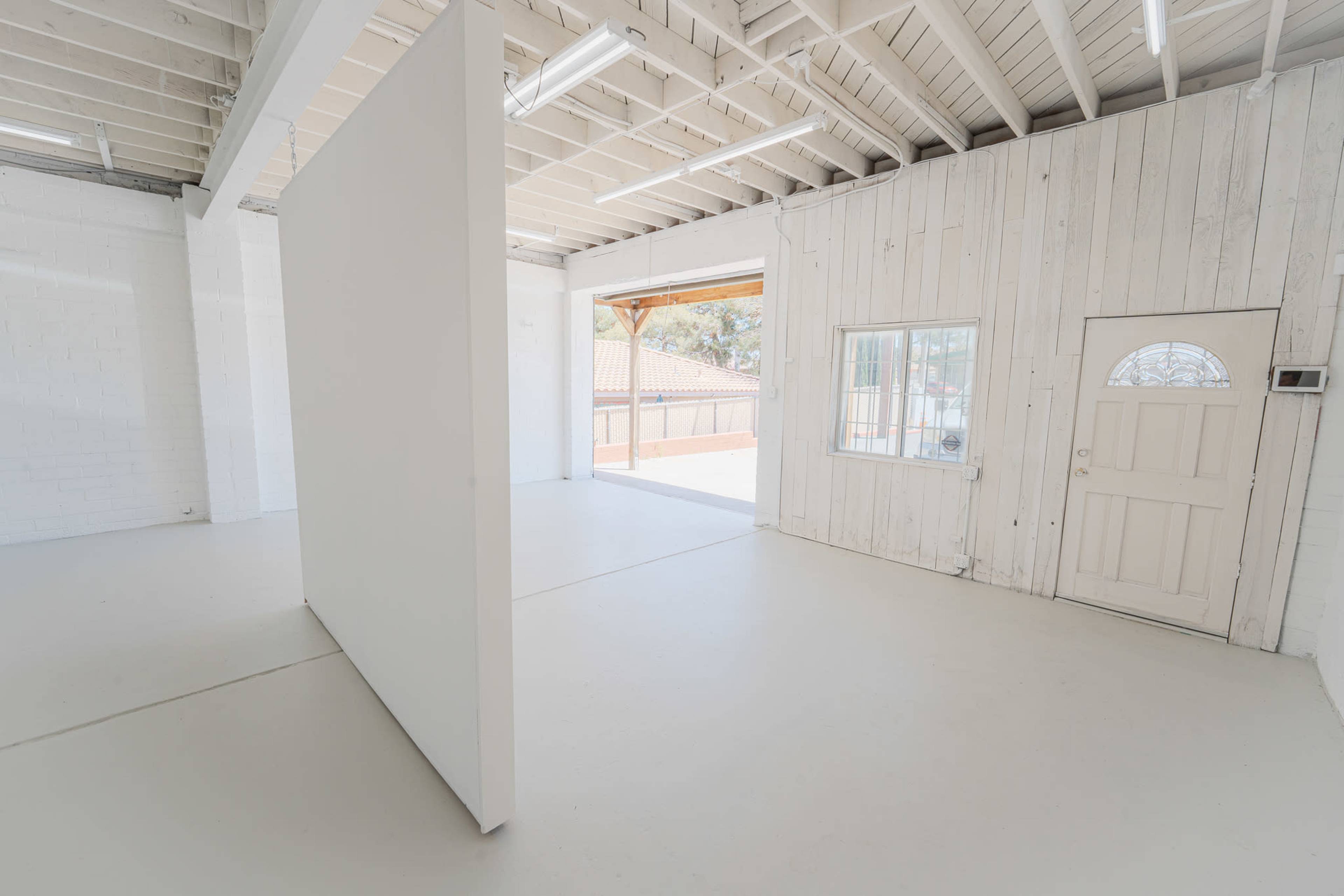The image shows a spacious, well-lit room with a white partition wall and an open doorway leading to an outdoor area.