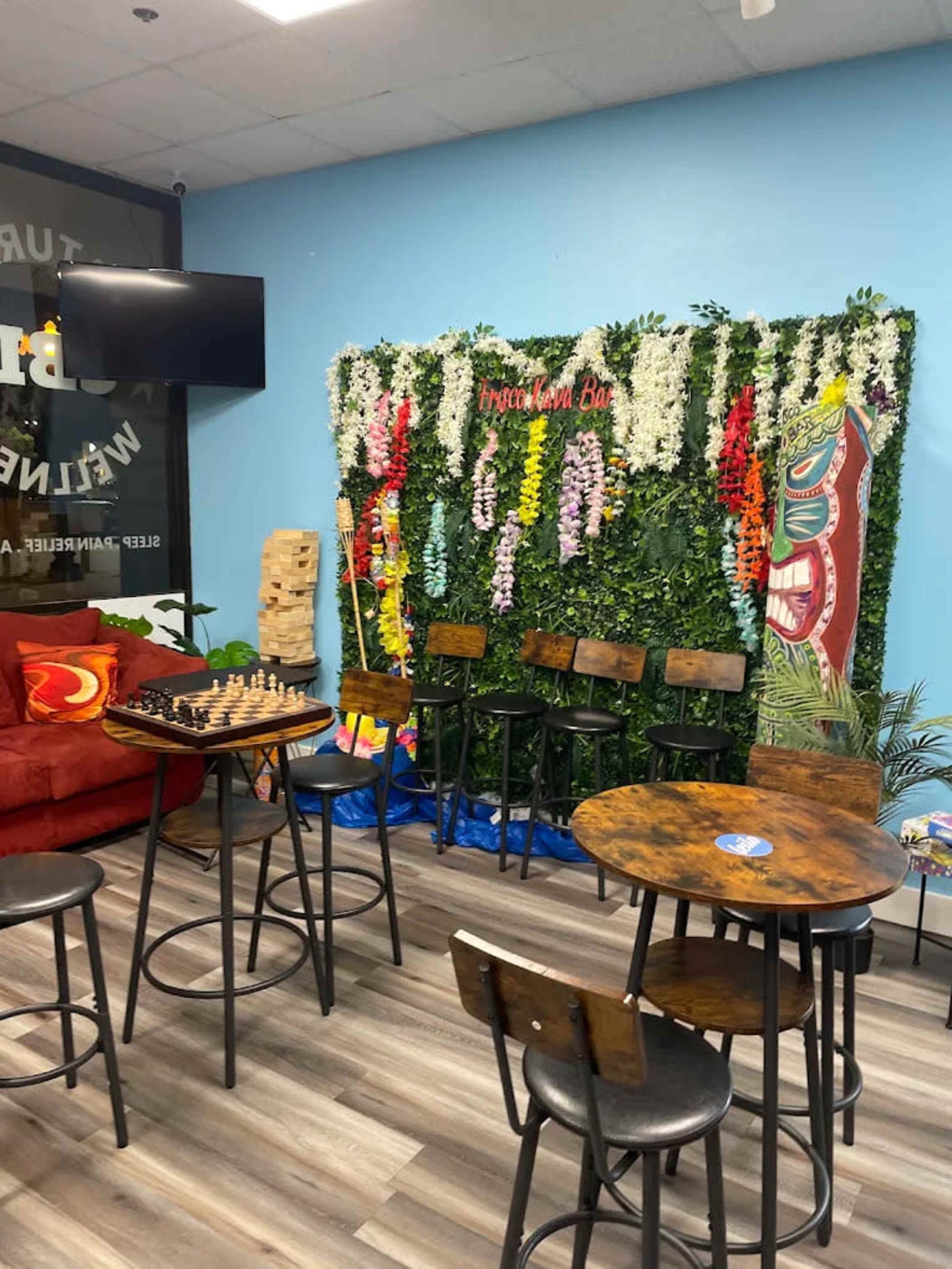 The image shows a cozy indoor space featuring a seating area with a red couch, a chessboard on a table, and bar stools in front of a decorative wall adorned with plants and colorful artwork.