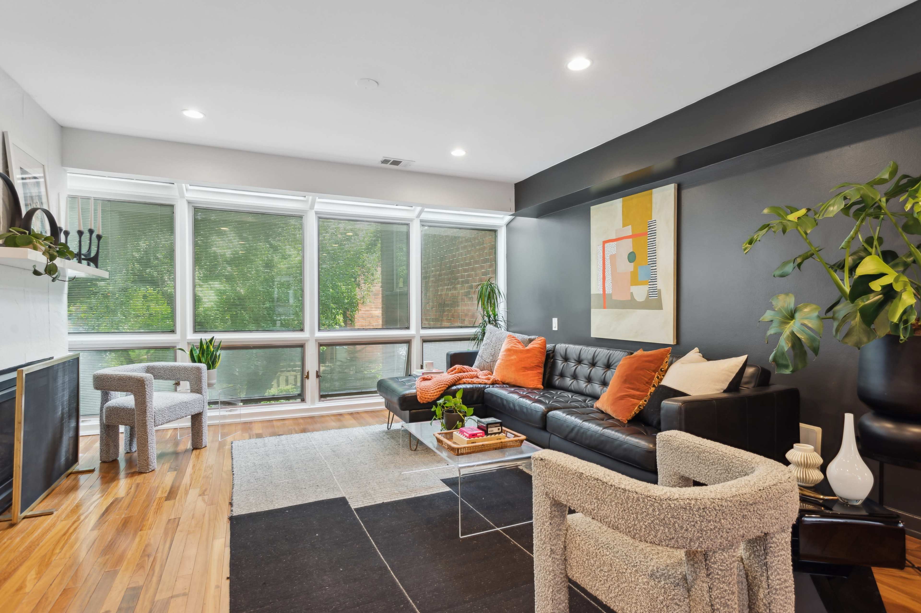 A modern living room with large windows, a black leather sofa, and a mix of decorative plants and furniture.