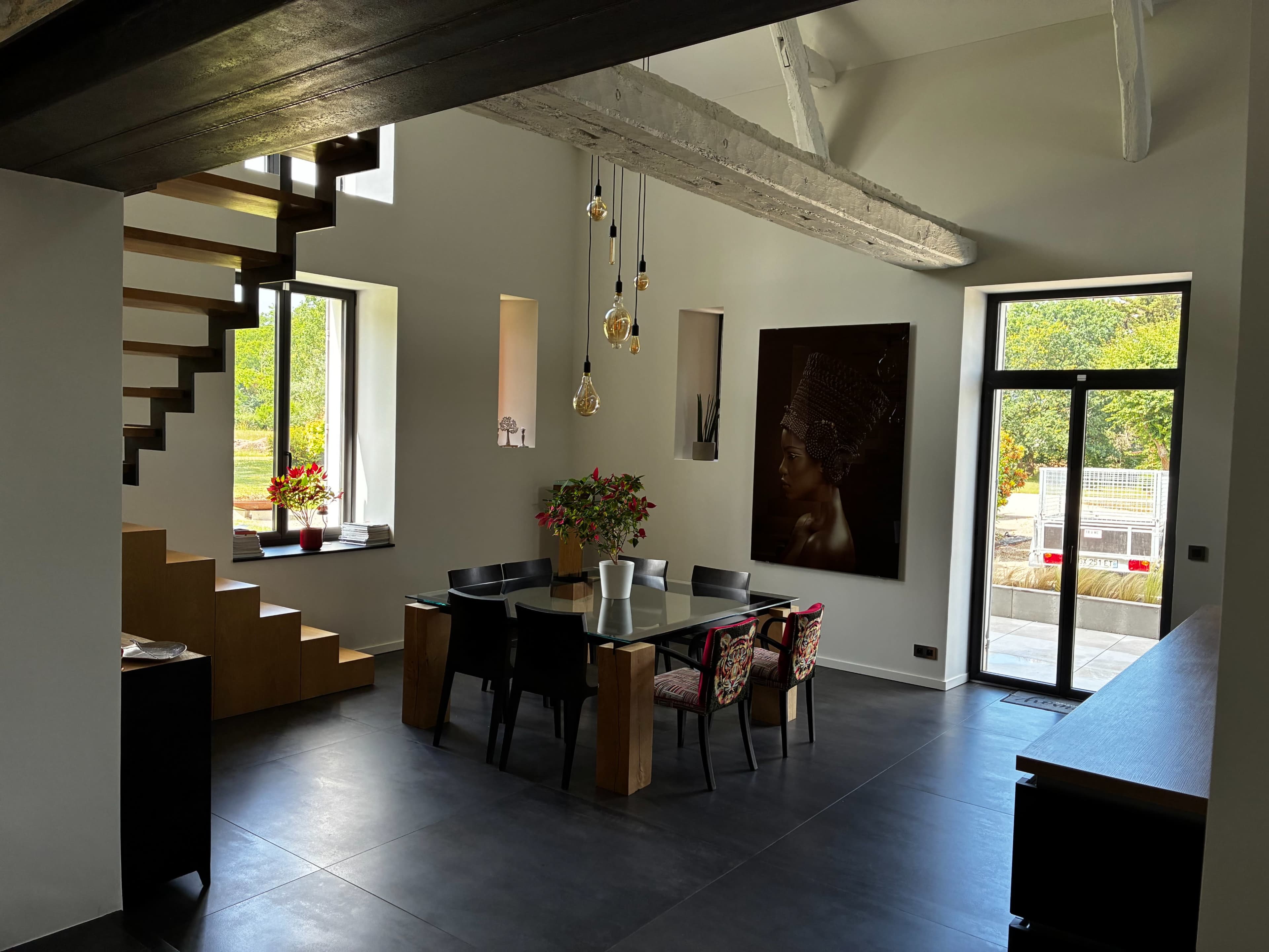 A modern dining area features a wooden table surrounded by colorful chairs, with a staircase leading to an upper level and large windows providing natural light.