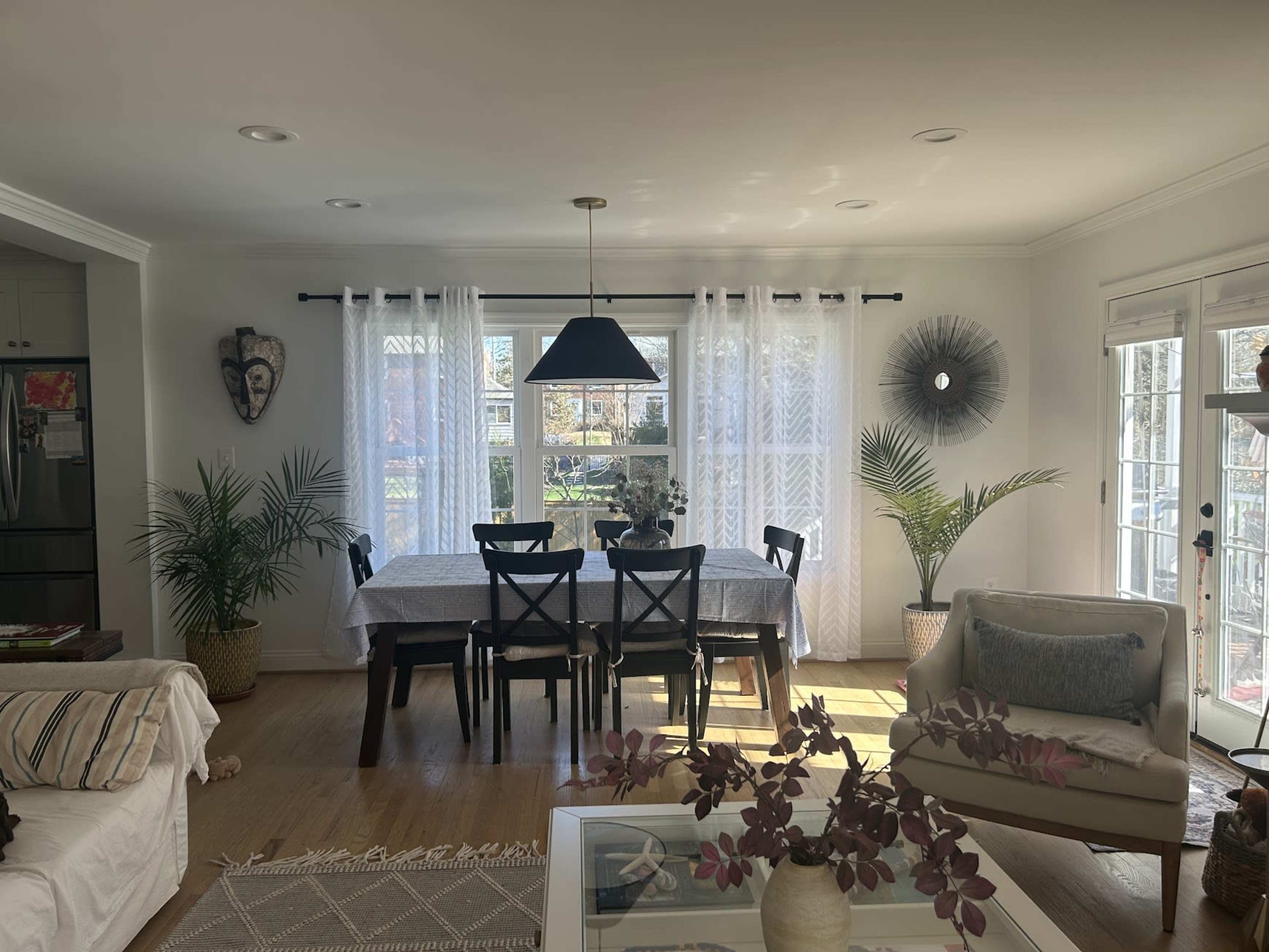 A dining area features a table surrounded by chairs, with large windows draped in sheer curtains letting in natural light, and various plants and decor elements complementing the space.