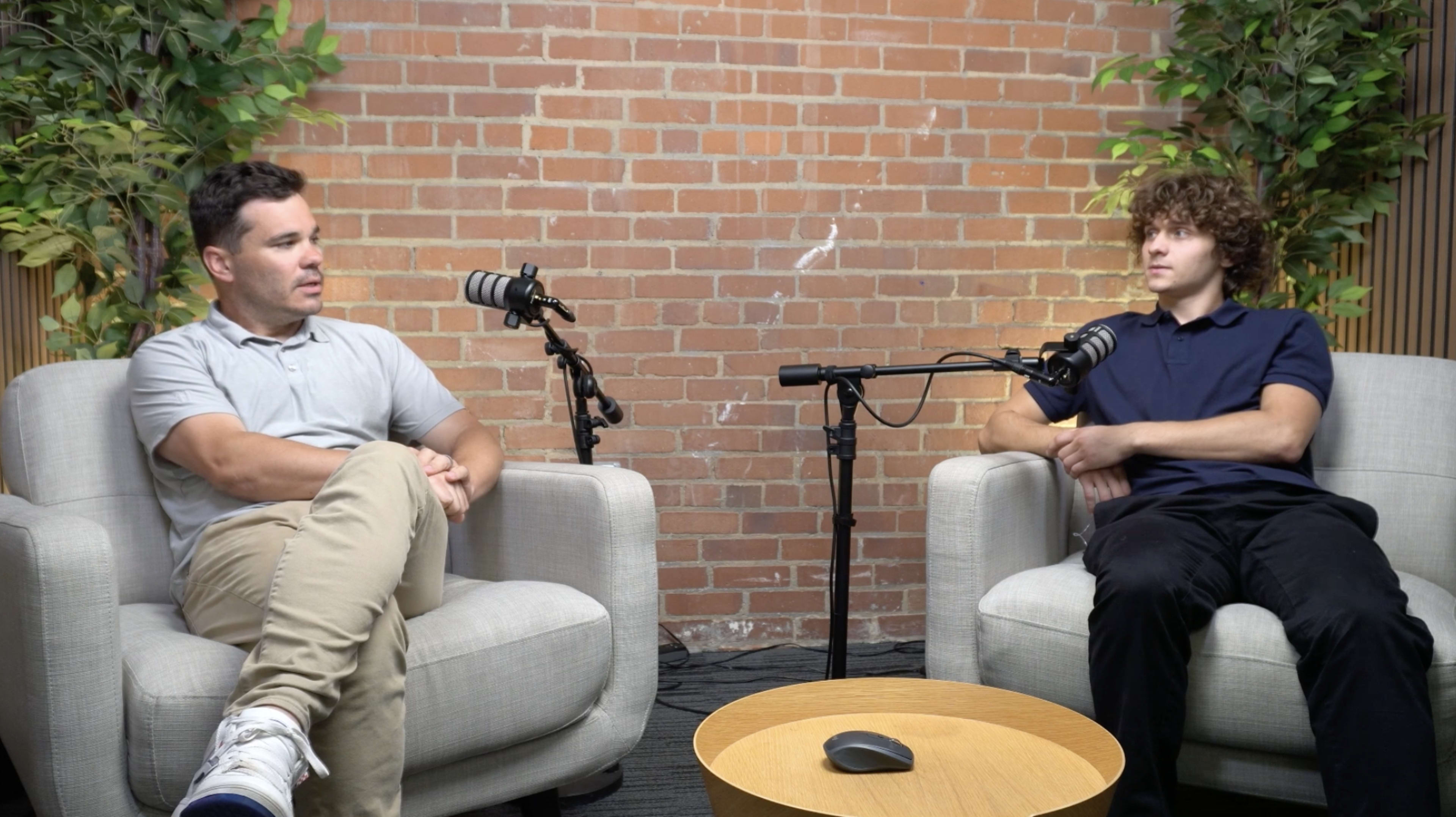 Two men are seated in armchairs facing each other in a room with a brick wall, flanked by plants, and equipped with microphones and a small round table in the center.