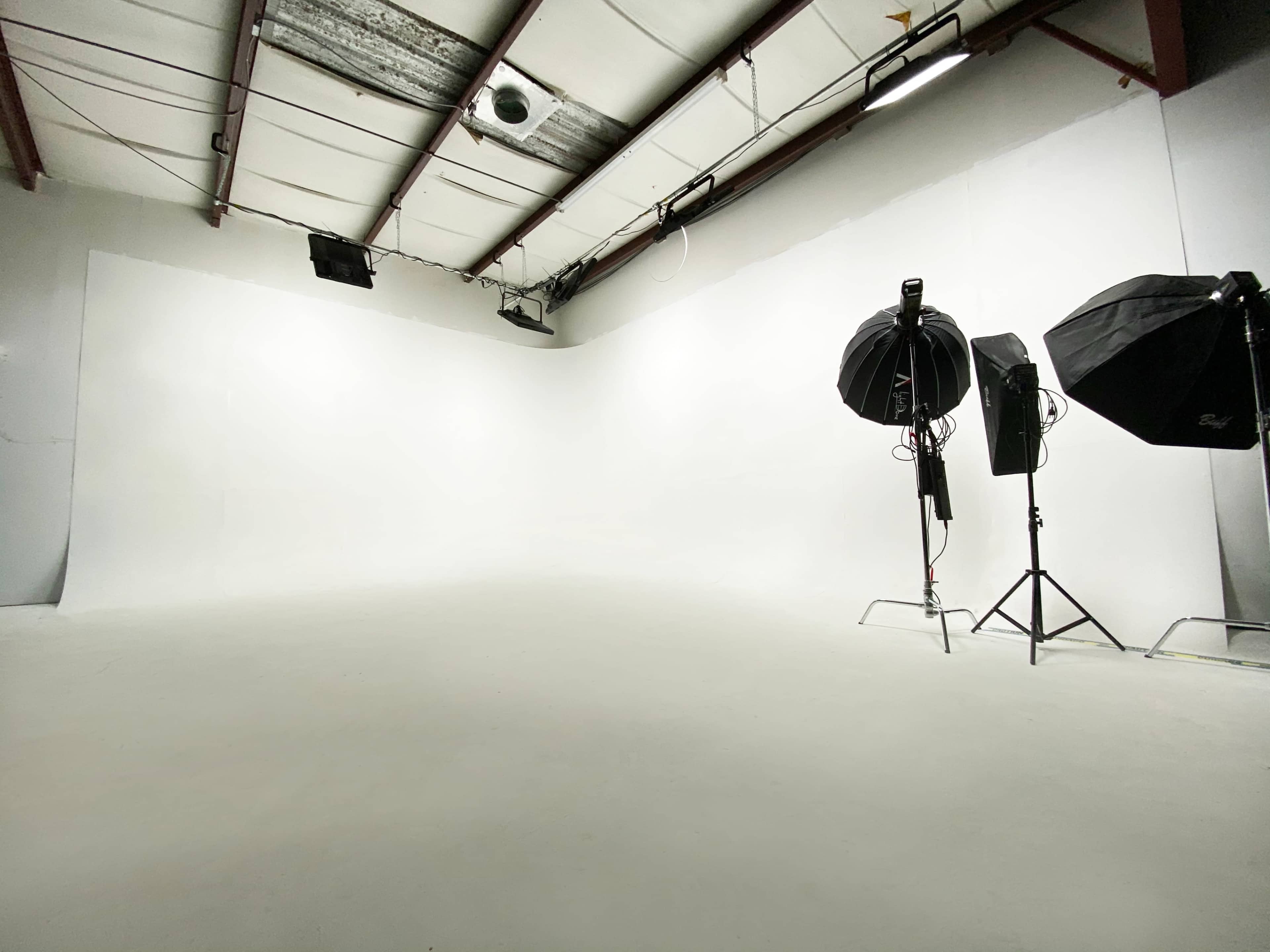 A spacious, empty photography studio with white walls and two lighting setups on stands.