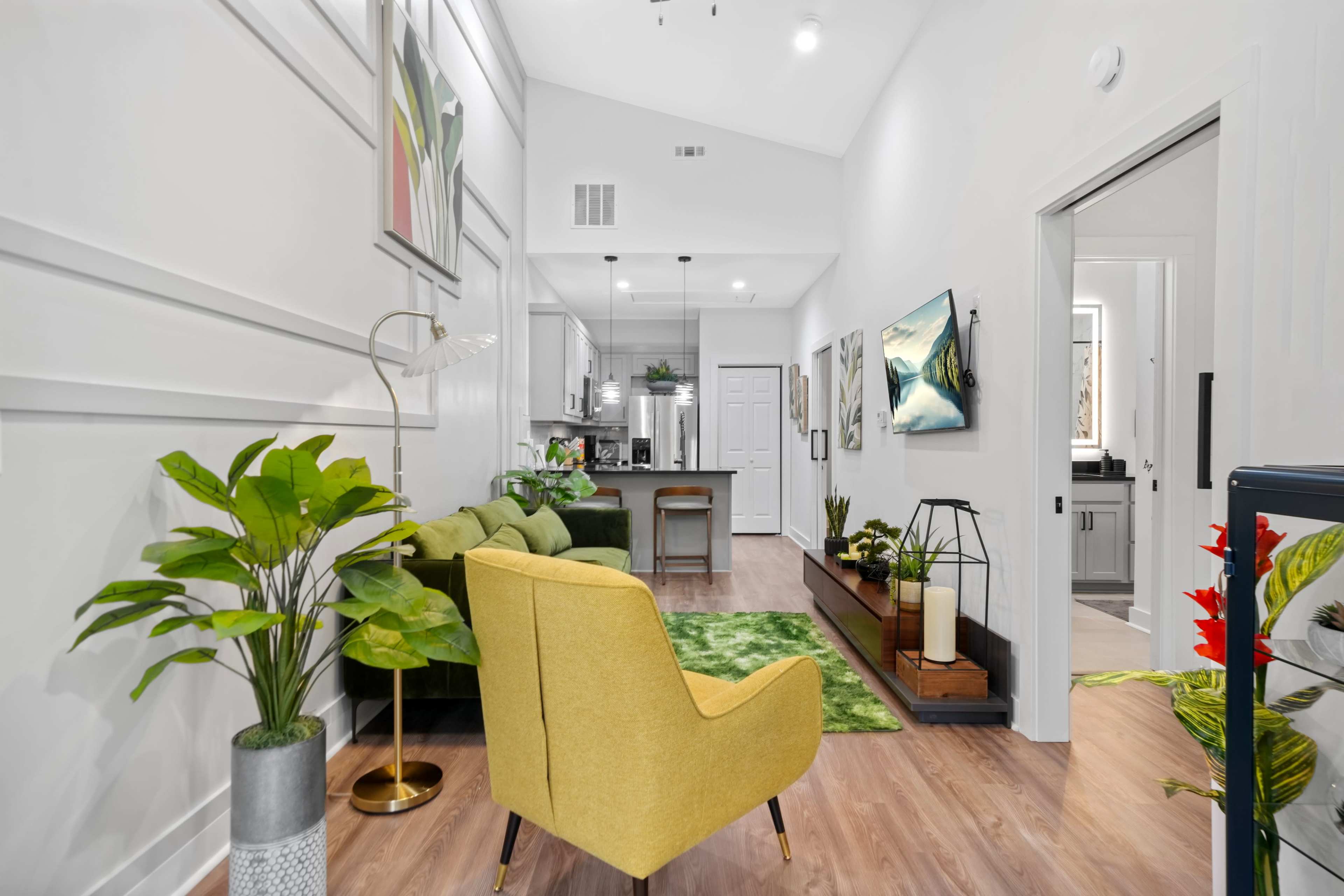 The image shows a modern living space with a green sofa, a yellow chair, and bright decorative elements, leading into a kitchen area.