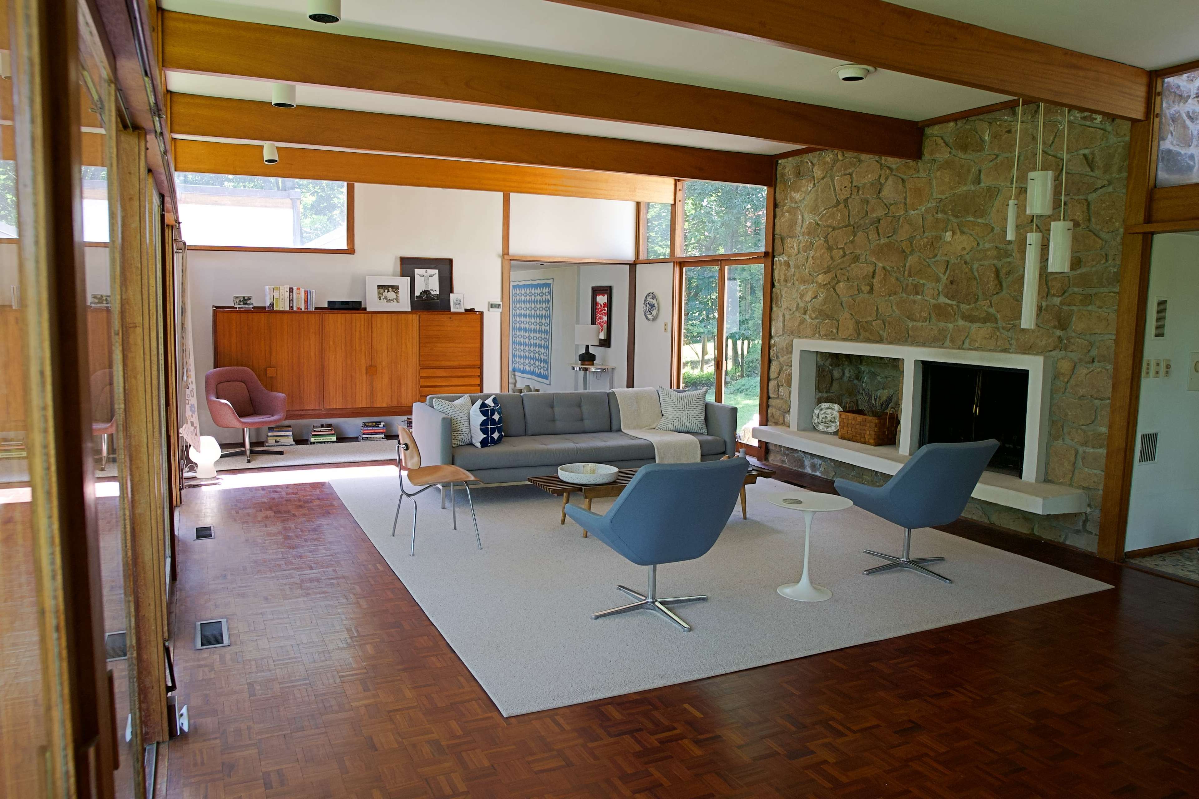 The image shows a modern living room with a stone fireplace, a gray sofa, two blue chairs, and large windows allowing natural light to enter.