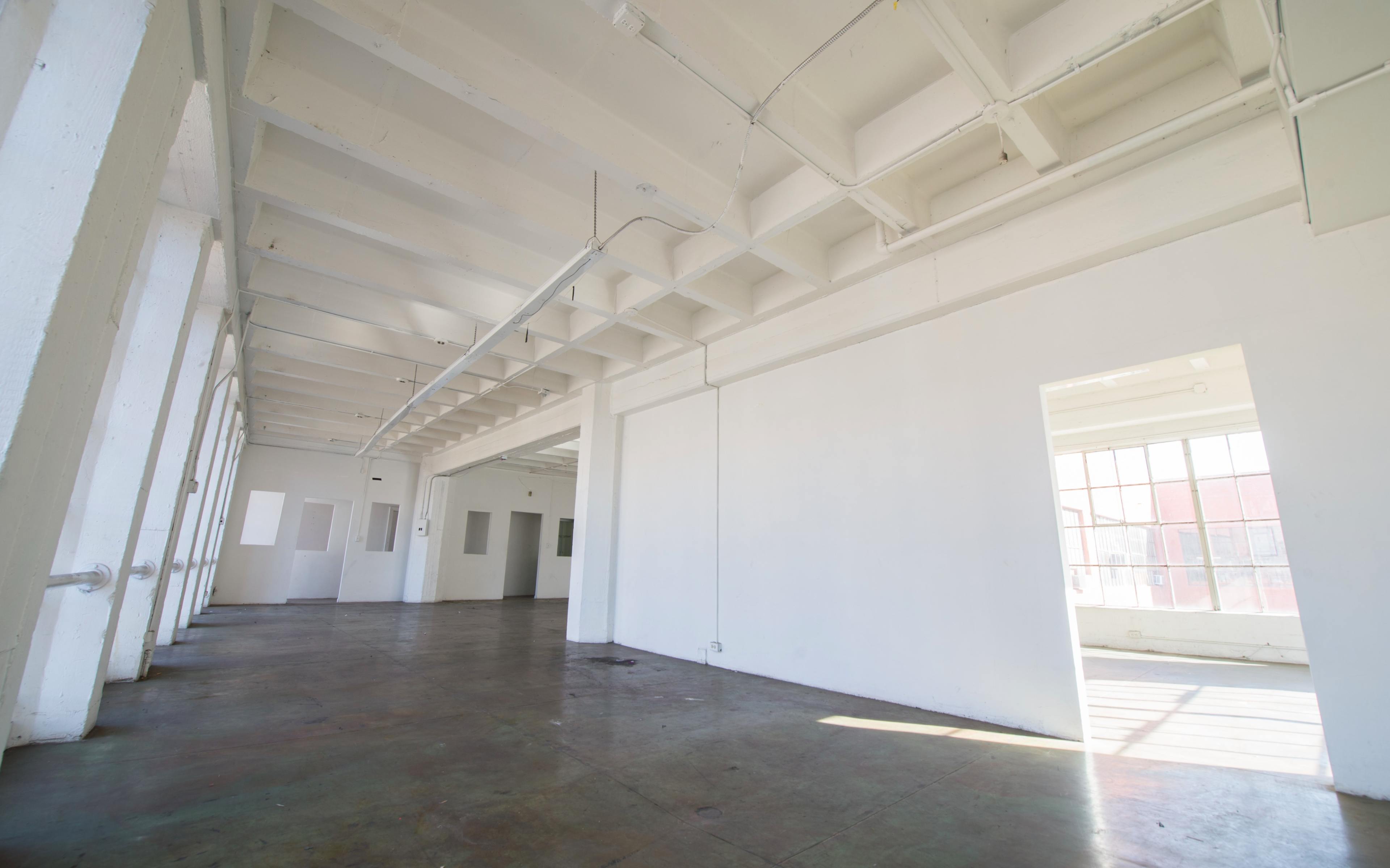 A spacious, empty room with white walls, high ceilings, and large windows letting in natural light.