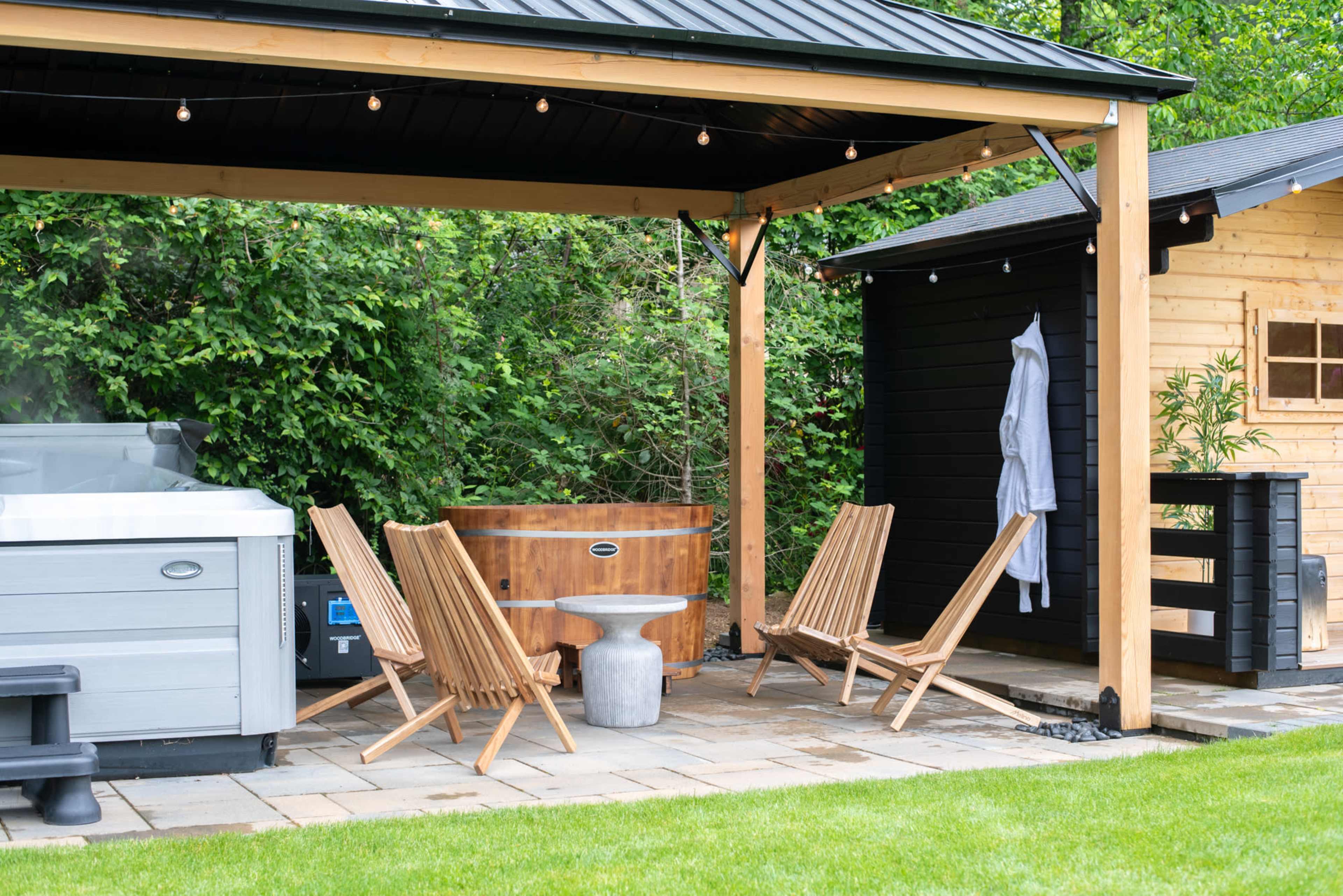 The image shows a backyard area with a hot tub and wooden chairs under a gazebo, alongside a small shed and green landscaping.