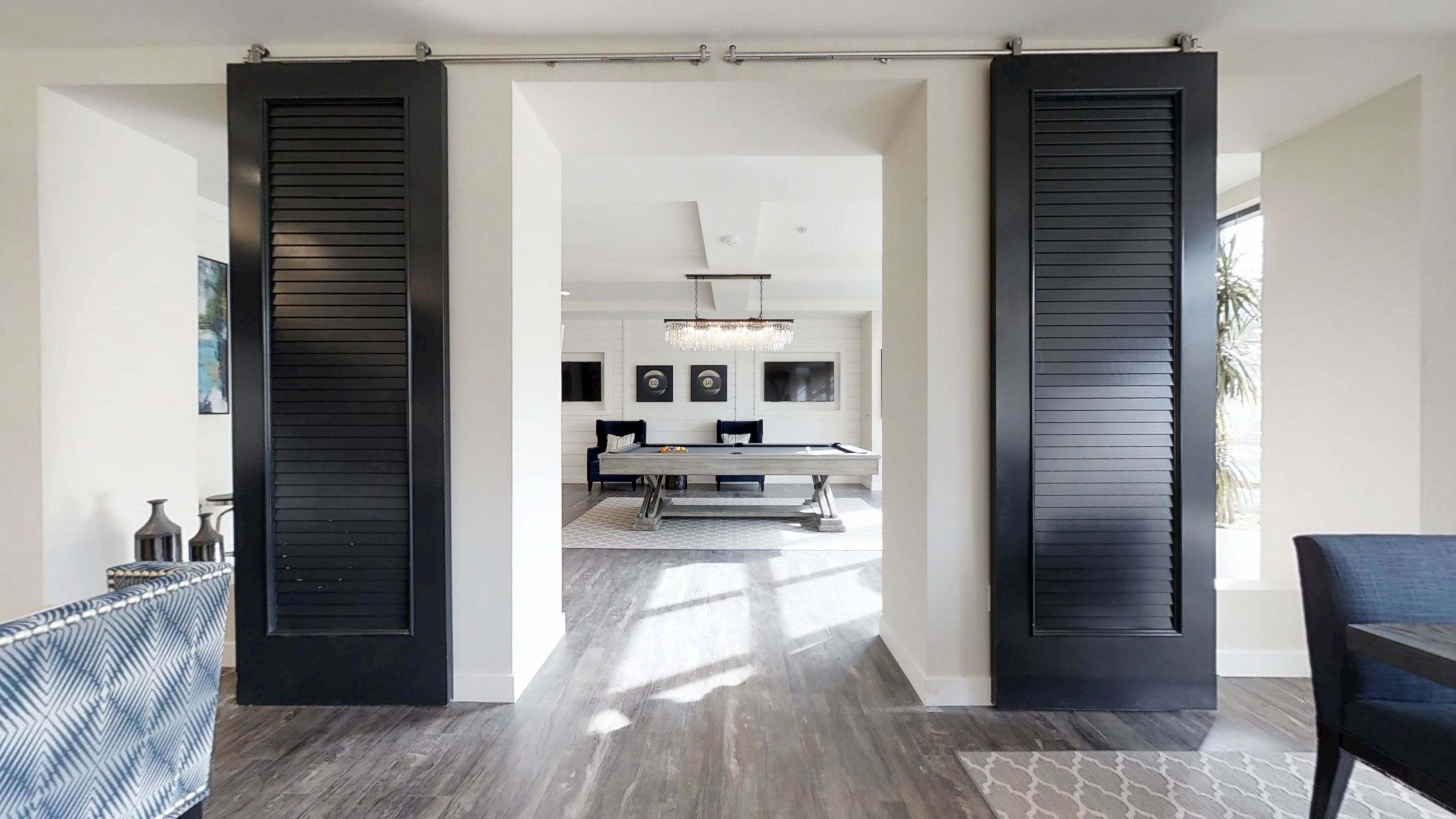 The image shows a modern interior space featuring black louvered doors opening to a well-lit area with a pool table and elegant decor.