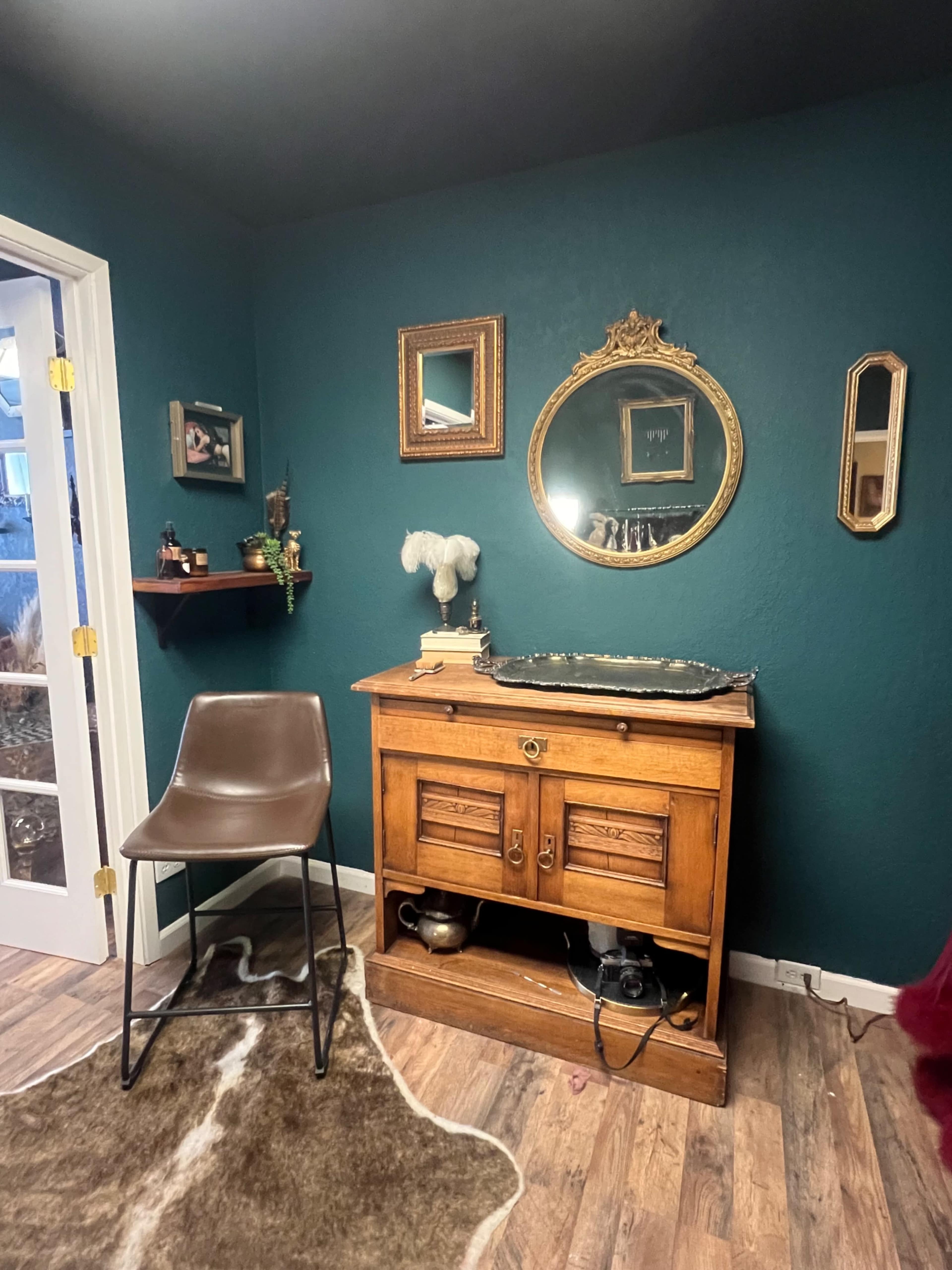 The image shows a wooden sideboard with a decorative mirror above it, accompanied by a chair and shelves on a dark teal wall.