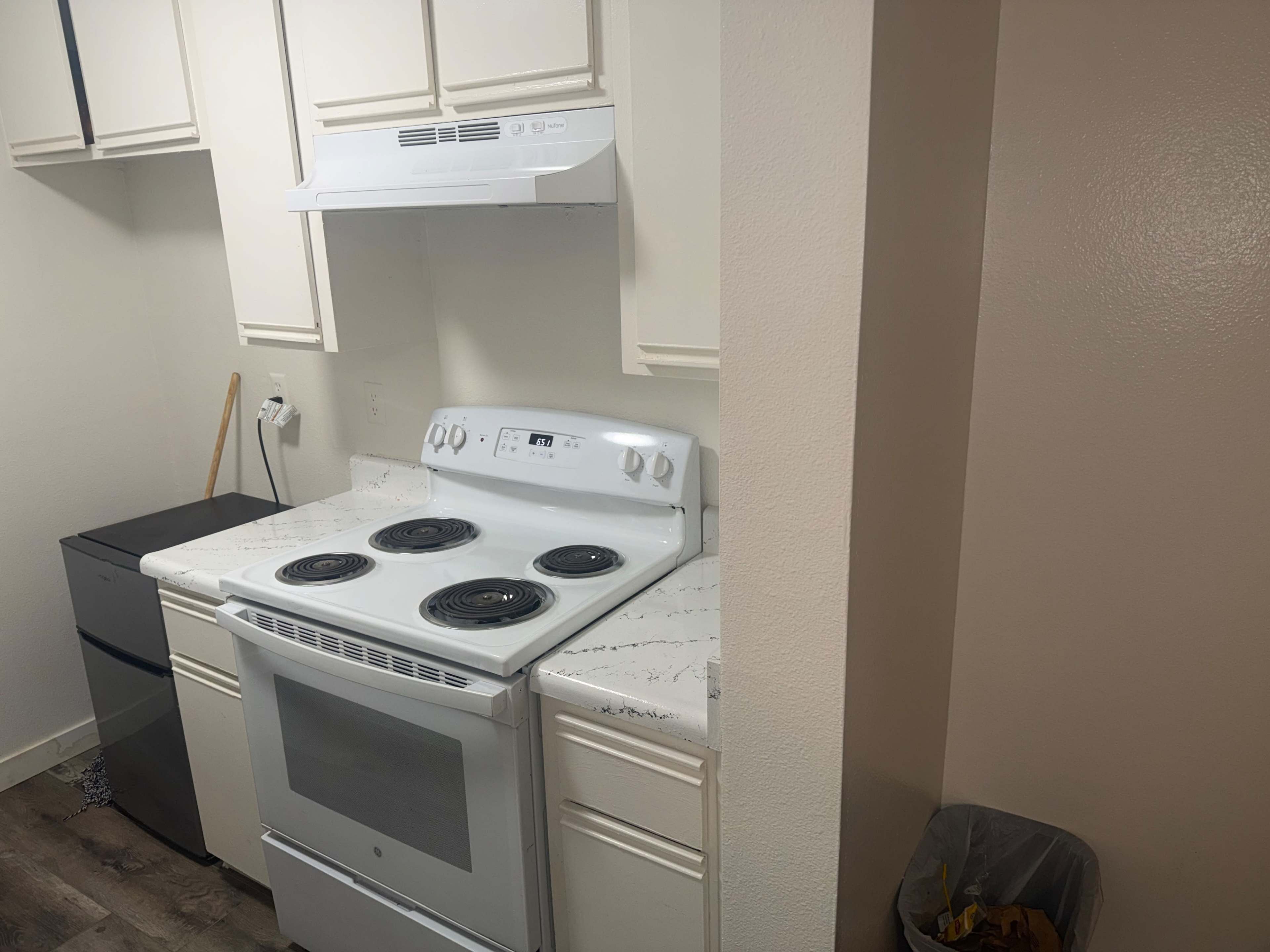 The image shows a kitchen corner featuring a white stove with four burners, a hood above it, and light-colored cabinets alongside a small refrigerator and a trash can.