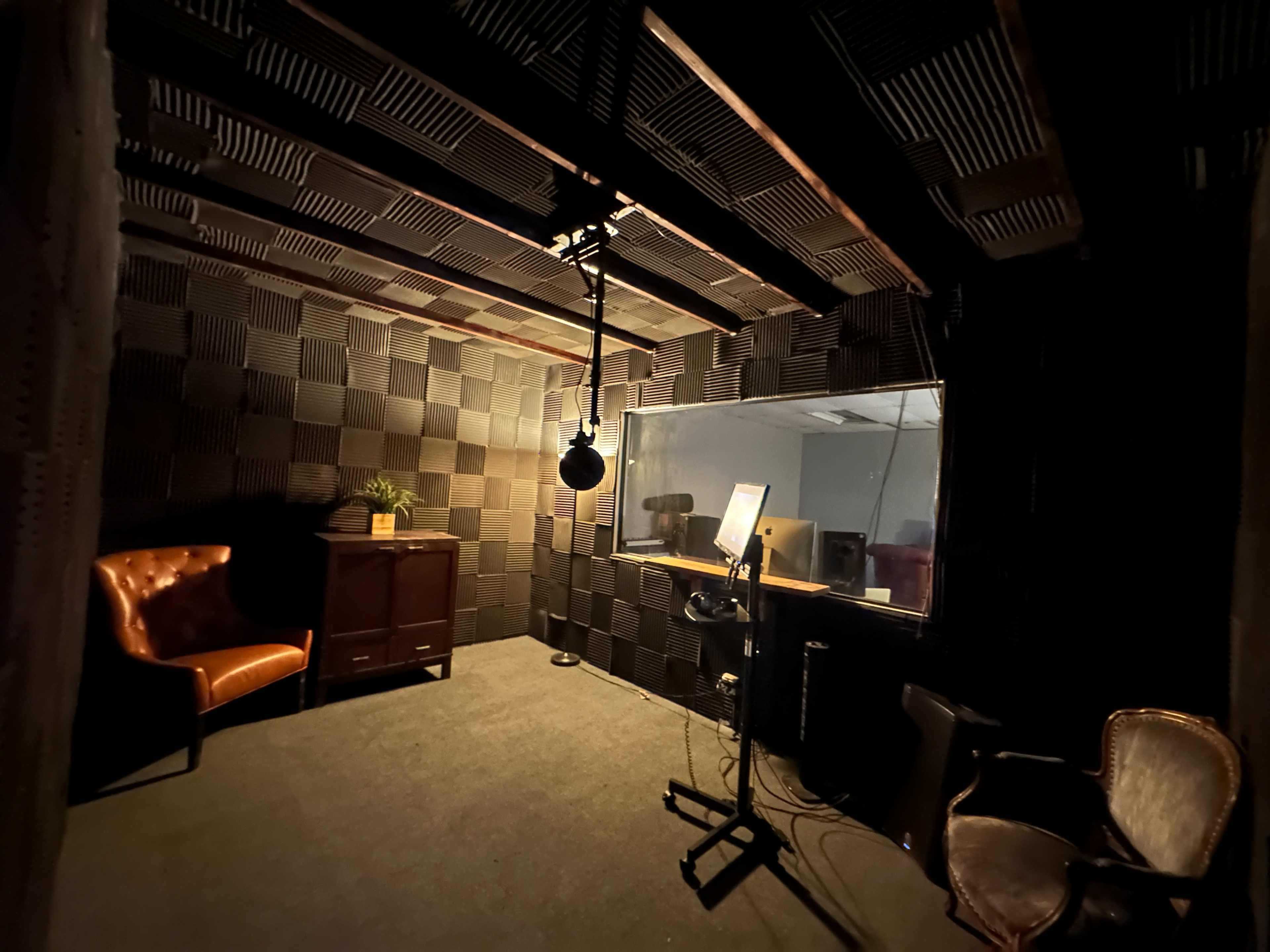 The image shows a soundproof recording studio with a microphone, a desk, and chairs, surrounded by acoustic panels.