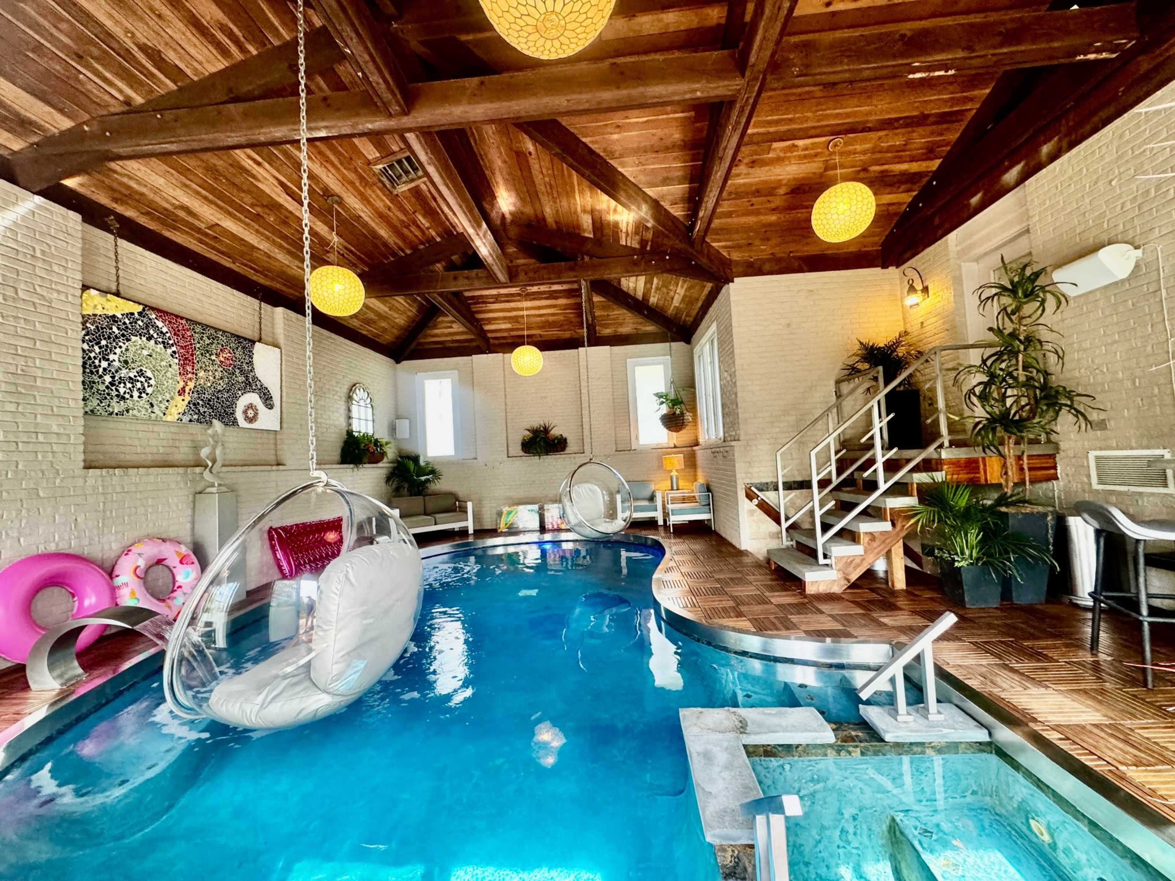 The image shows an indoor pool area featuring a hanging chair, a jacuzzi, and various plants under a wooden ceiling with decorative lighting.